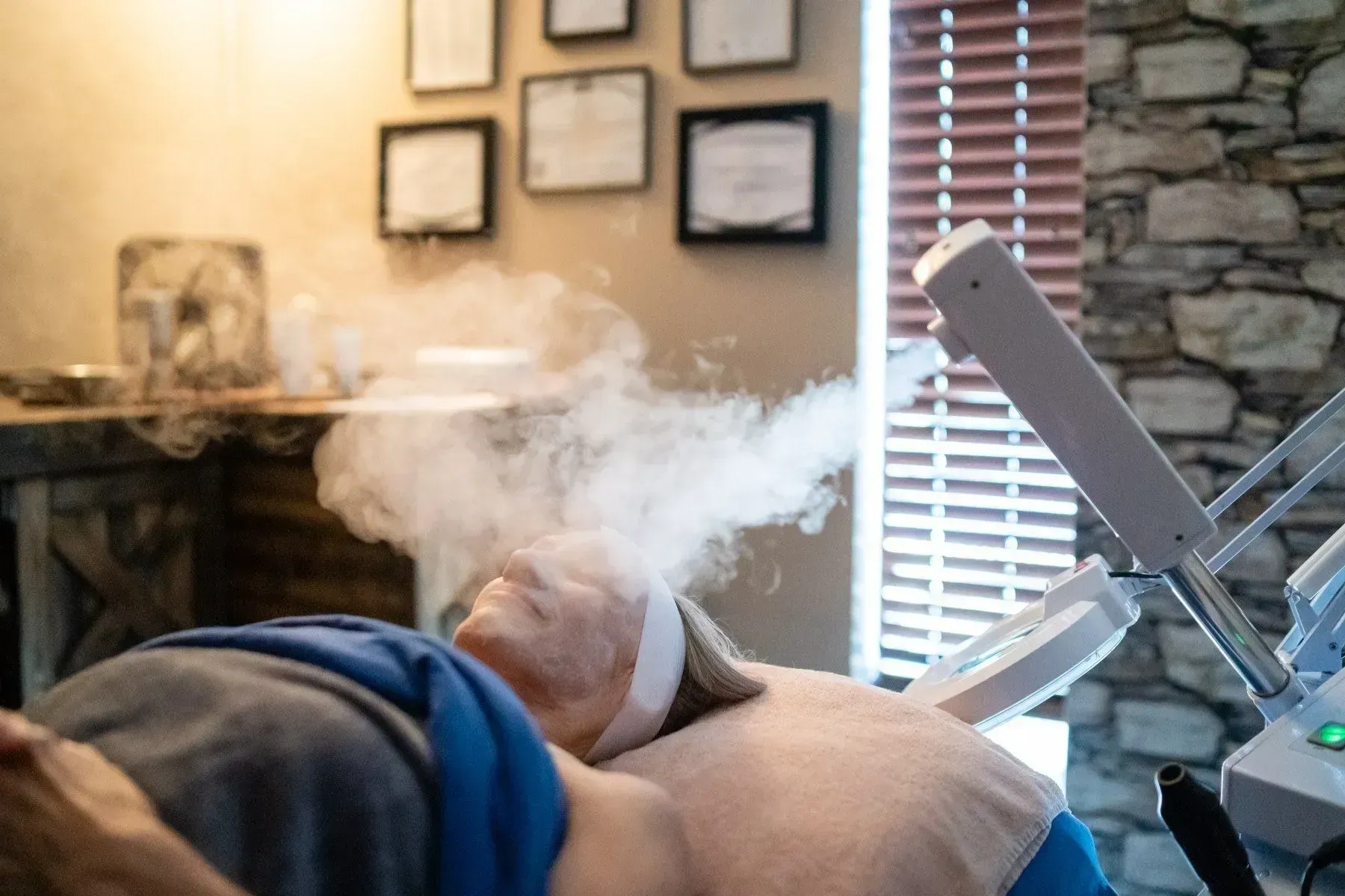 Woman receiving facial steam treatment in a spa. White steam surrounds her face; beige walls, certificates.