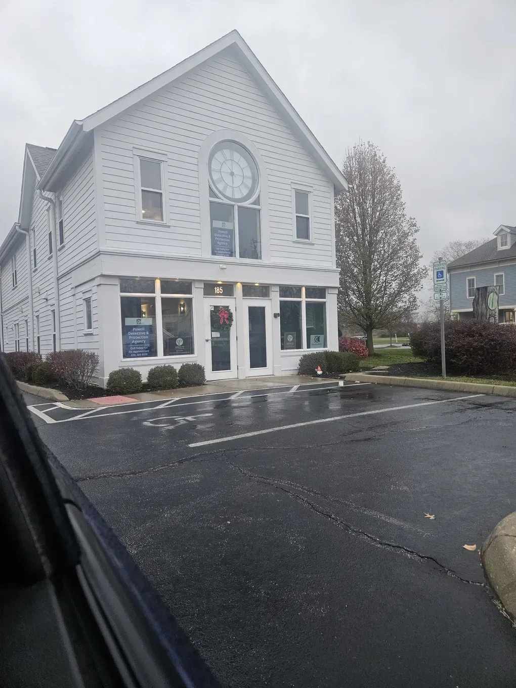 White building with a large clock on its front, two stories, glass doors, gray parking lot, and cloudy sky.