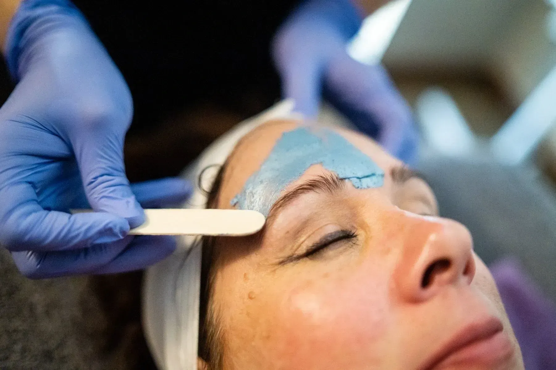 Woman getting eyebrow wax; blue wax on brow area; spa setting, closed eyes, gloved hands, pale skin.