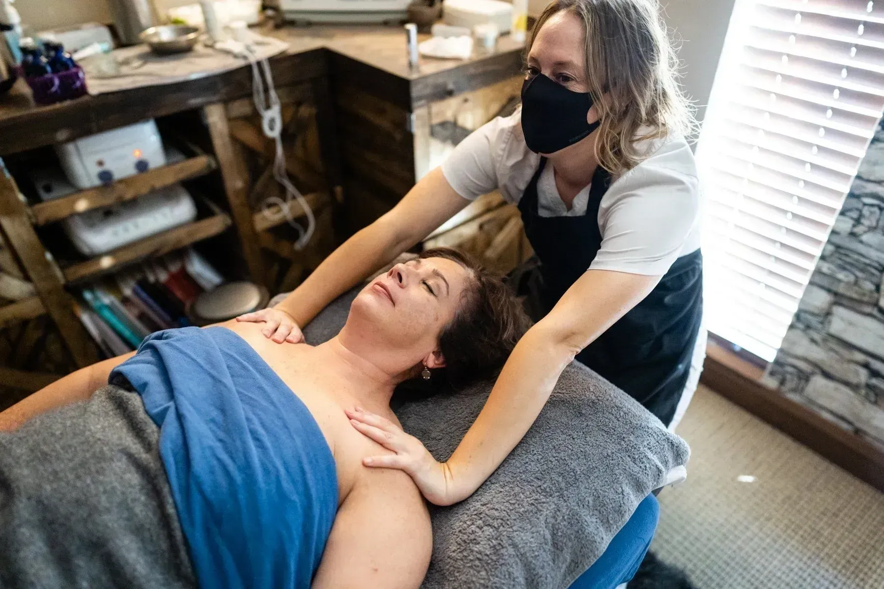 A woman in mask gives a massage to another woman lying face up on a table.