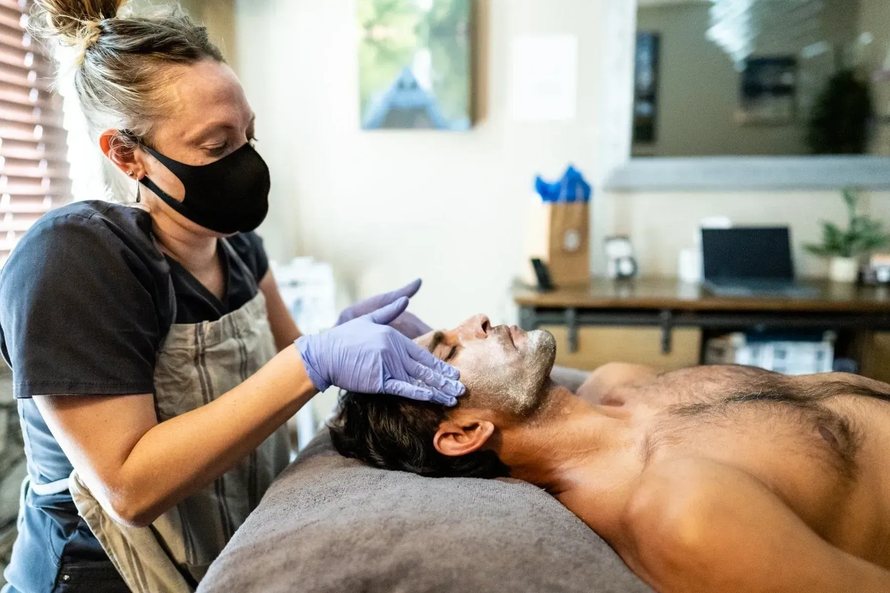Woman in mask and gloves giving a facial to a shirtless man lying down; indoor setting.