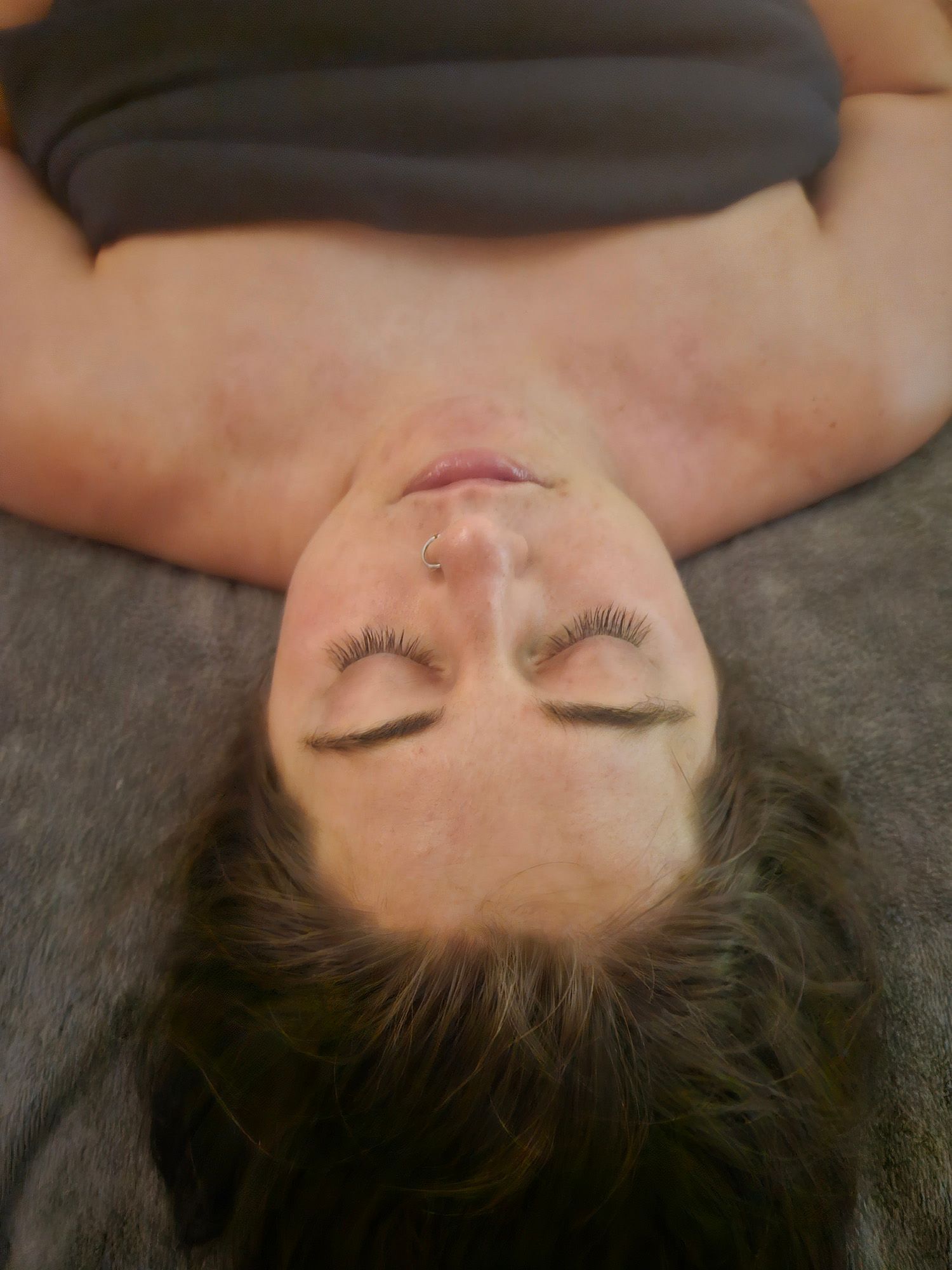 Woman with closed eyes, lying face up; shoulders and chest visible, on a gray surface.