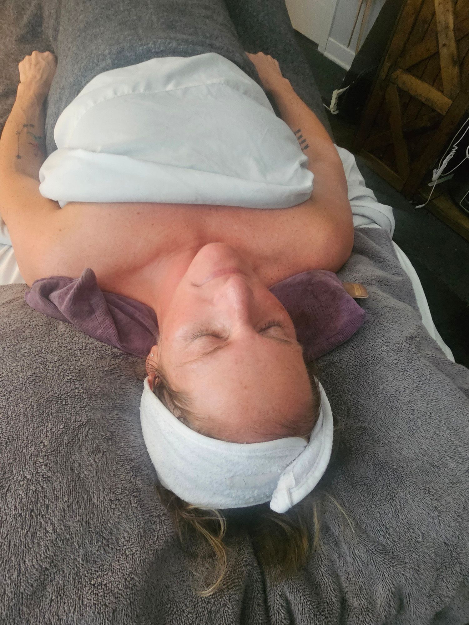 Woman reclining, eyes closed, wearing a headband and face towel, on a massage table.