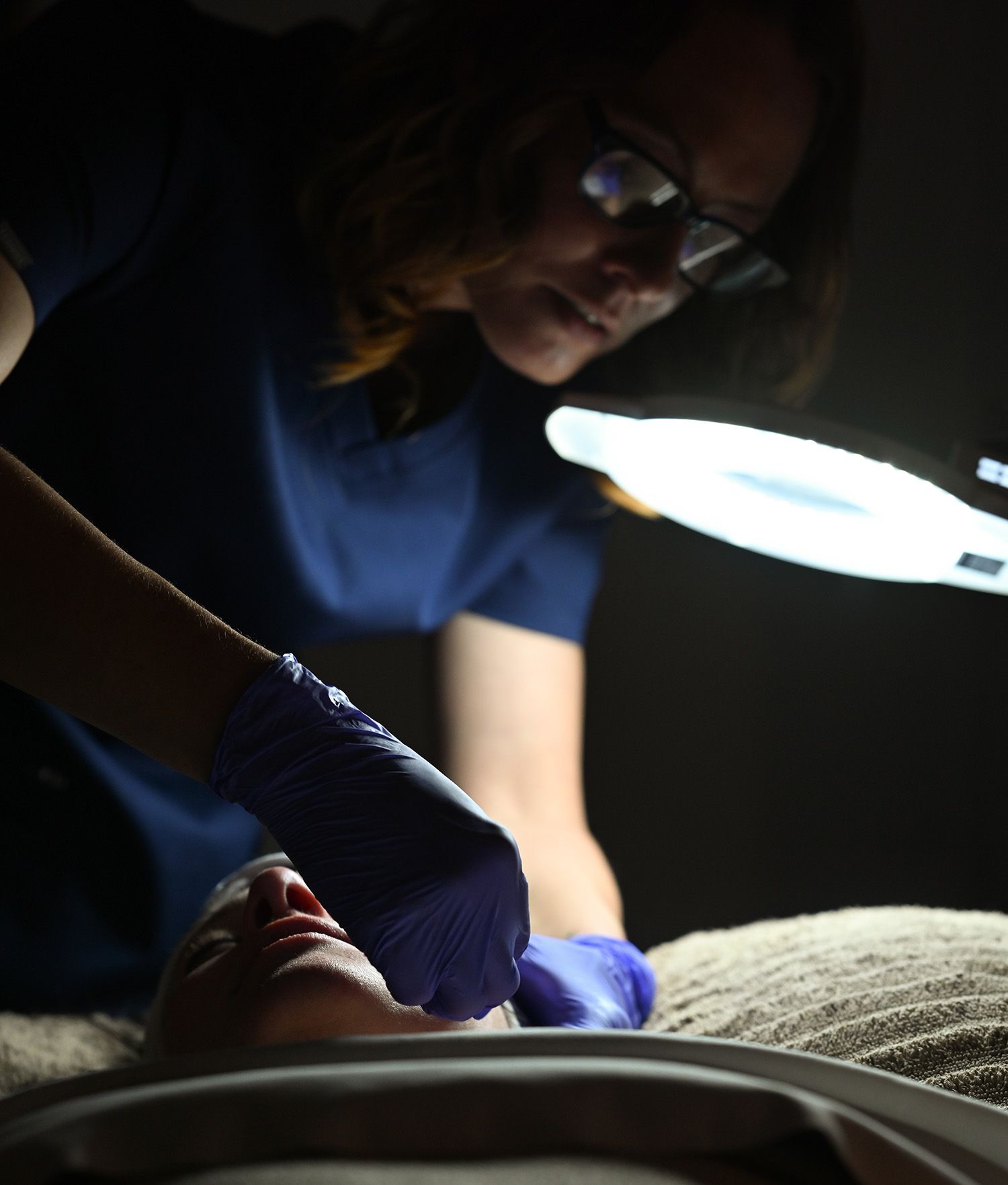 Aesthetician in blue scrubs performing a facial procedure on a client under bright light.