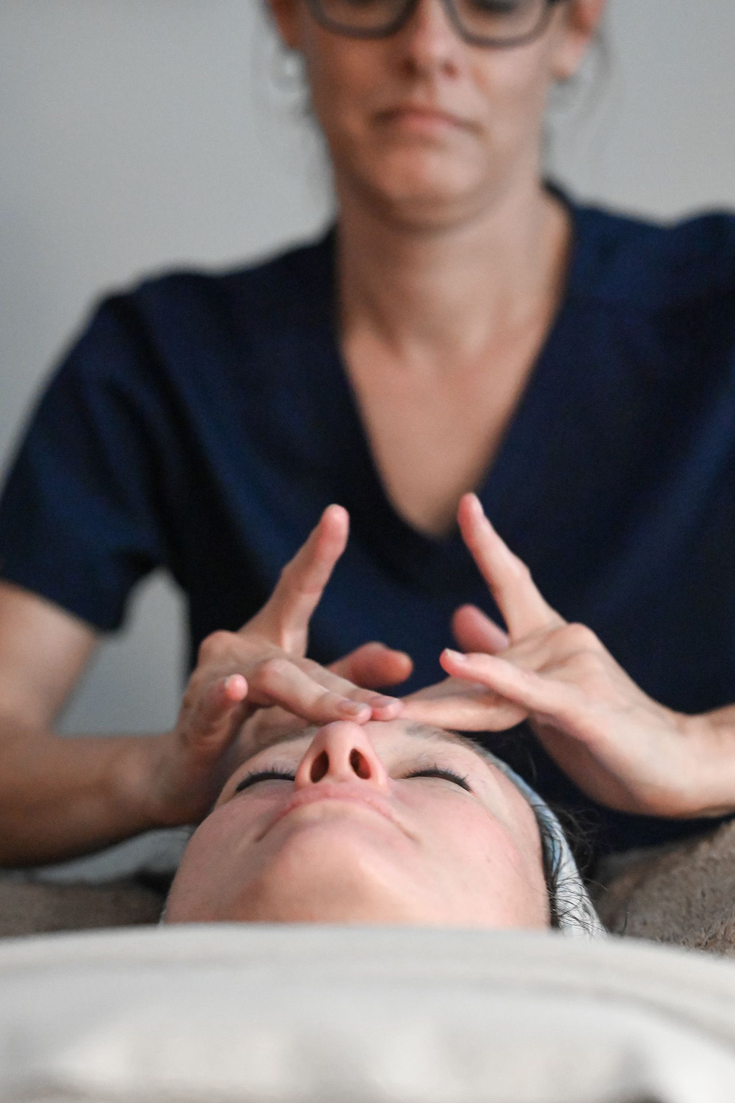 Woman receiving a facial massage; therapist's hands on forehead, blue scrub top, indoors.