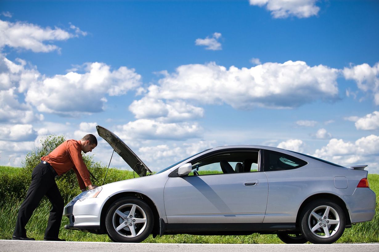 Person pushing a white car with its hood open on a roadside under a blue sky