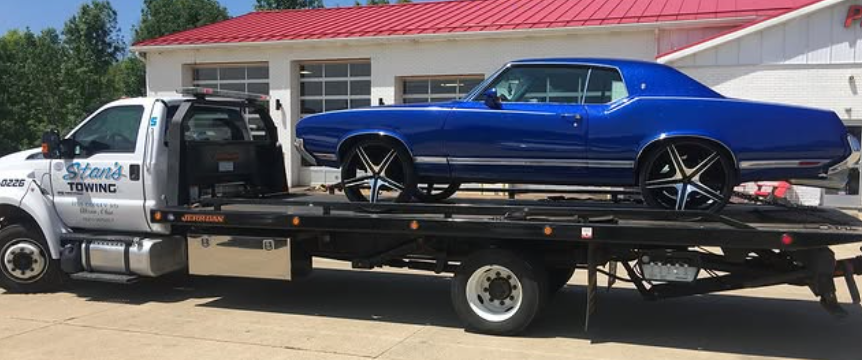 Tow truck hauling a blue classic car on a flatbed in front of a building