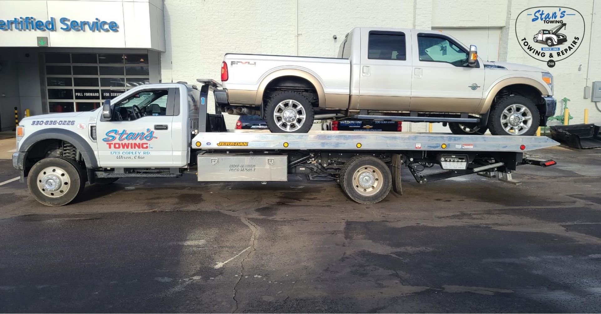 Two beige pickup trucks on a flatbed tow truck outside an auto service shop.