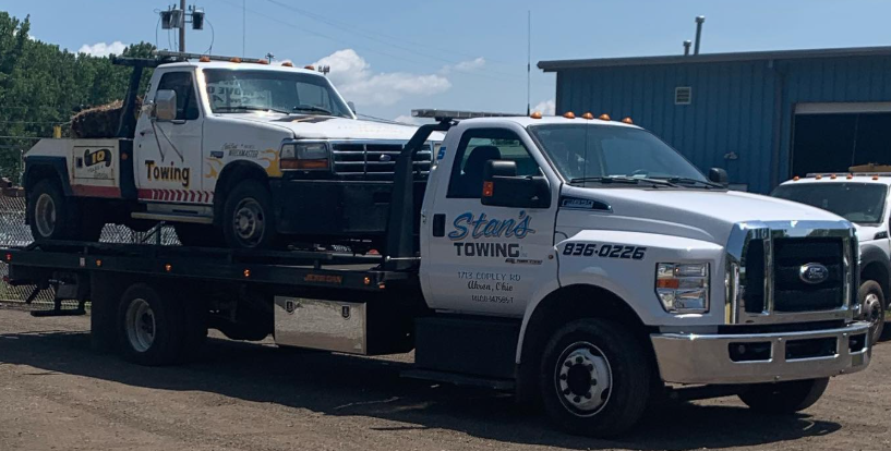 Tow truck carrying a white pickup and sedan in a sunny lot outside a blue building