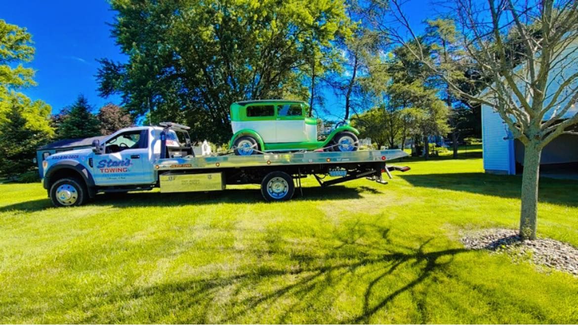 Tow truck hauling a green vintage car on a flatbed in a sunny grassy yard