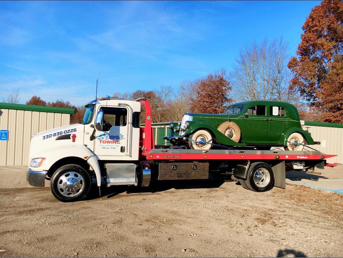 Tow truck carrying a green vintage car on a flatbed in a sunny outdoor lot