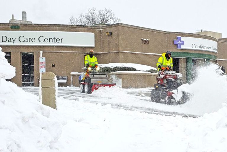 Two people in neon vests using snowblowers in front of a daycare center and a healthcare building.