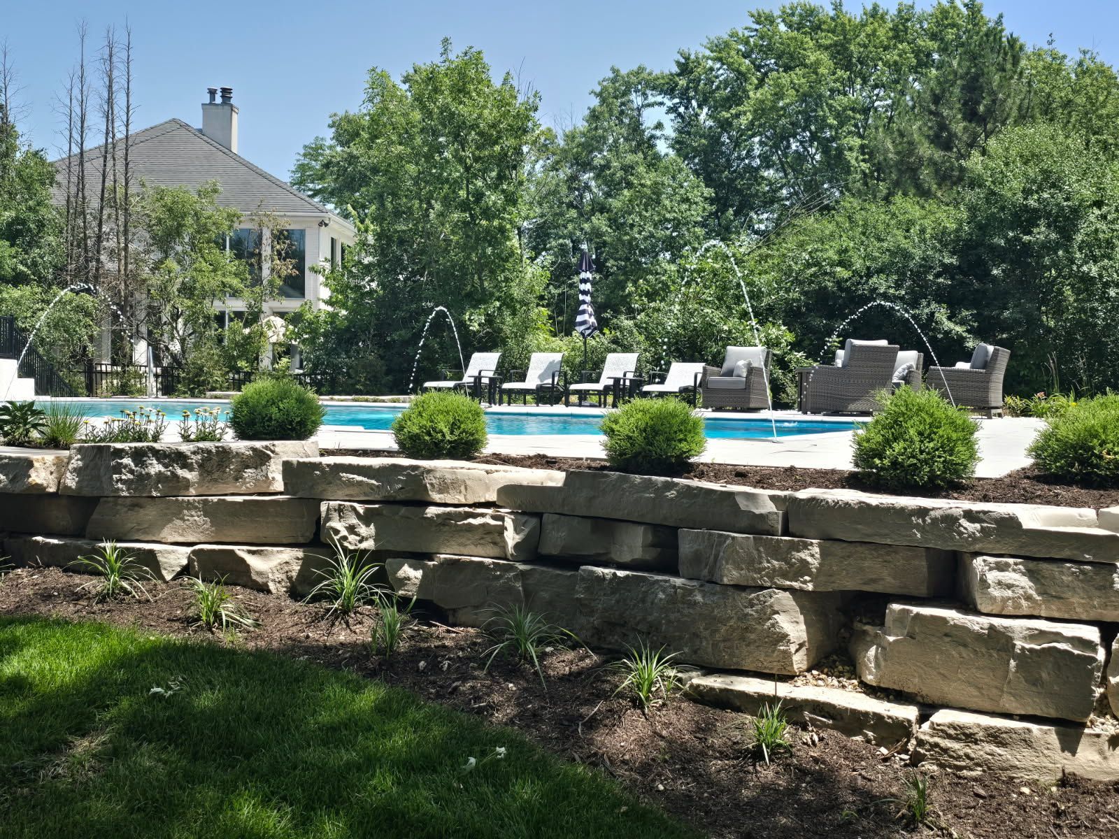 Stone patio with a swimming pool, lounge chairs, and trees in a sunny backyard garden.