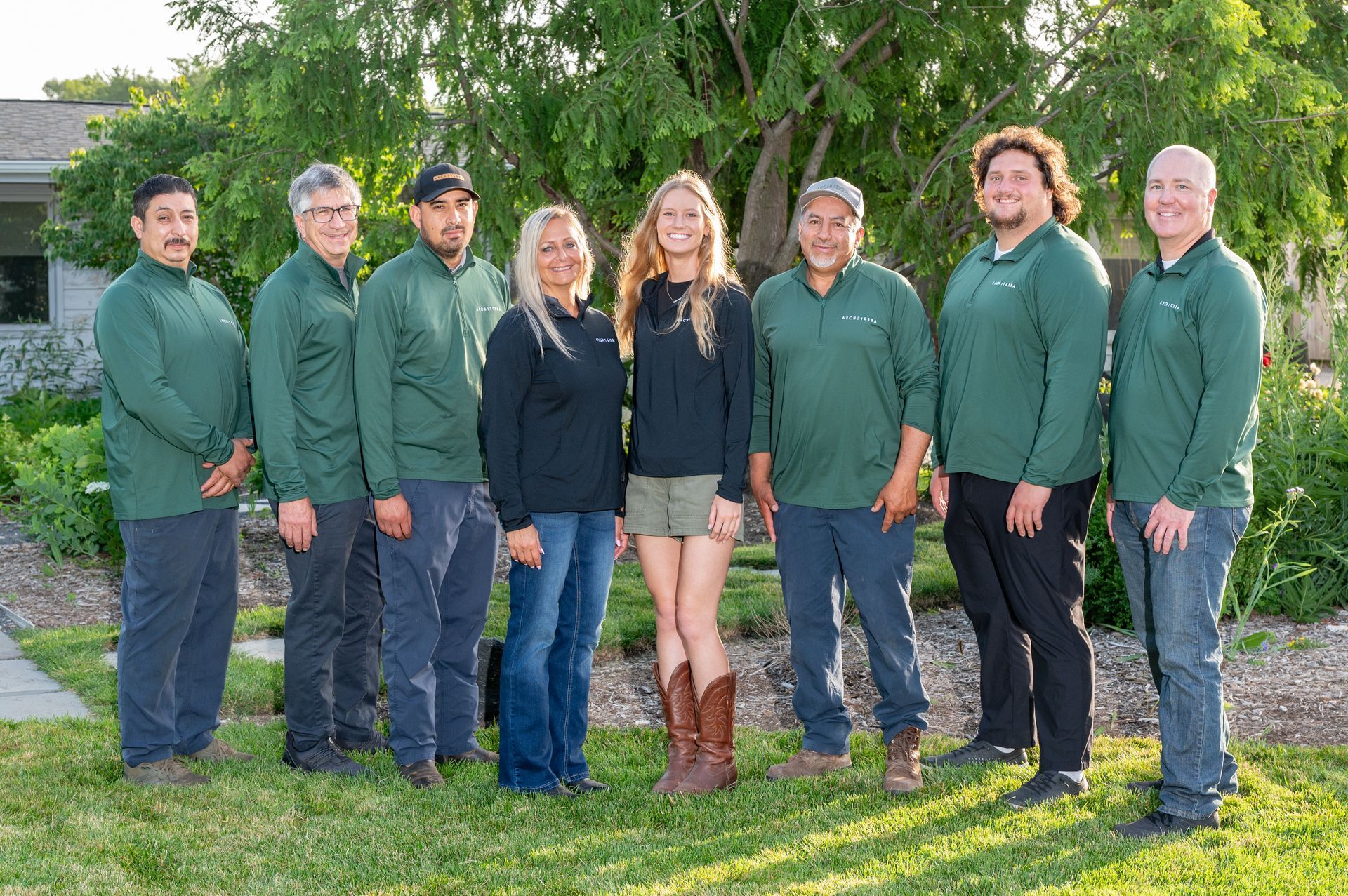 Eight people standing outdoors in matching green jackets, smiling on a sunny lawn.