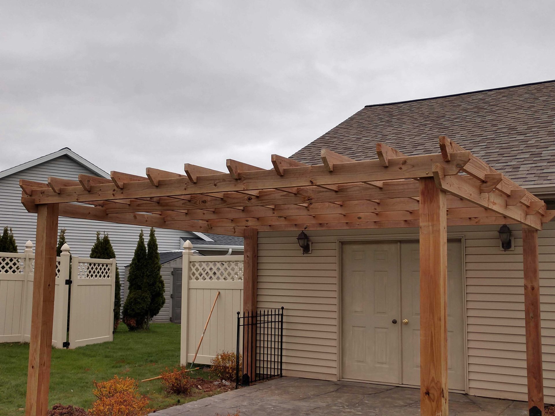 Wooden pergola attached to a light-colored building, set on a patio, with a white fence and cloudy sky.