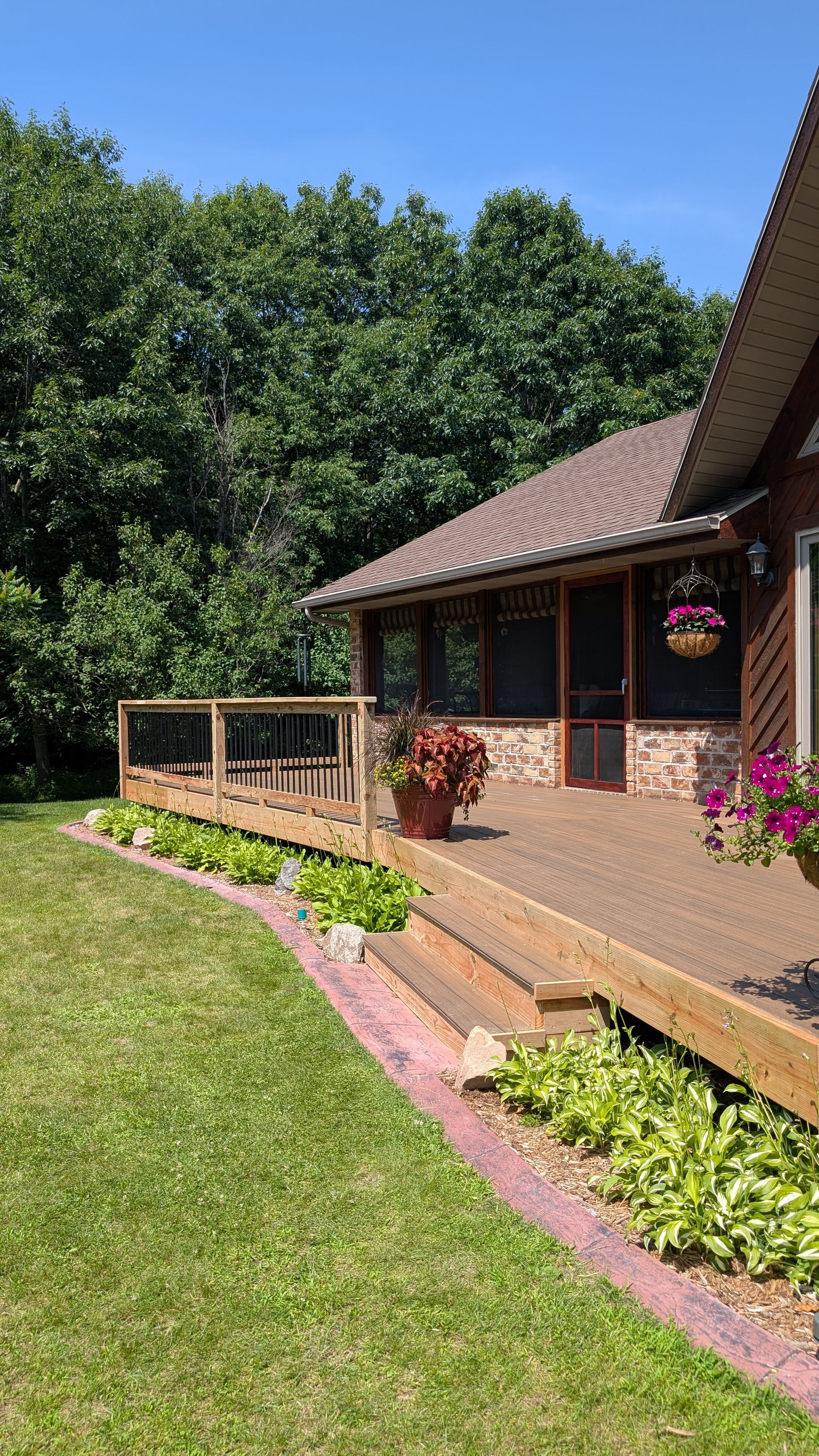 Brick house with a wooden porch, flower beds, and a green lawn beside trees under a blue sky