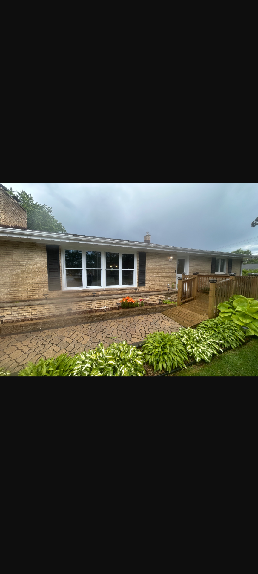 Single-story brick house with white-framed windows, wet front walkway, and greenery in the foreground.