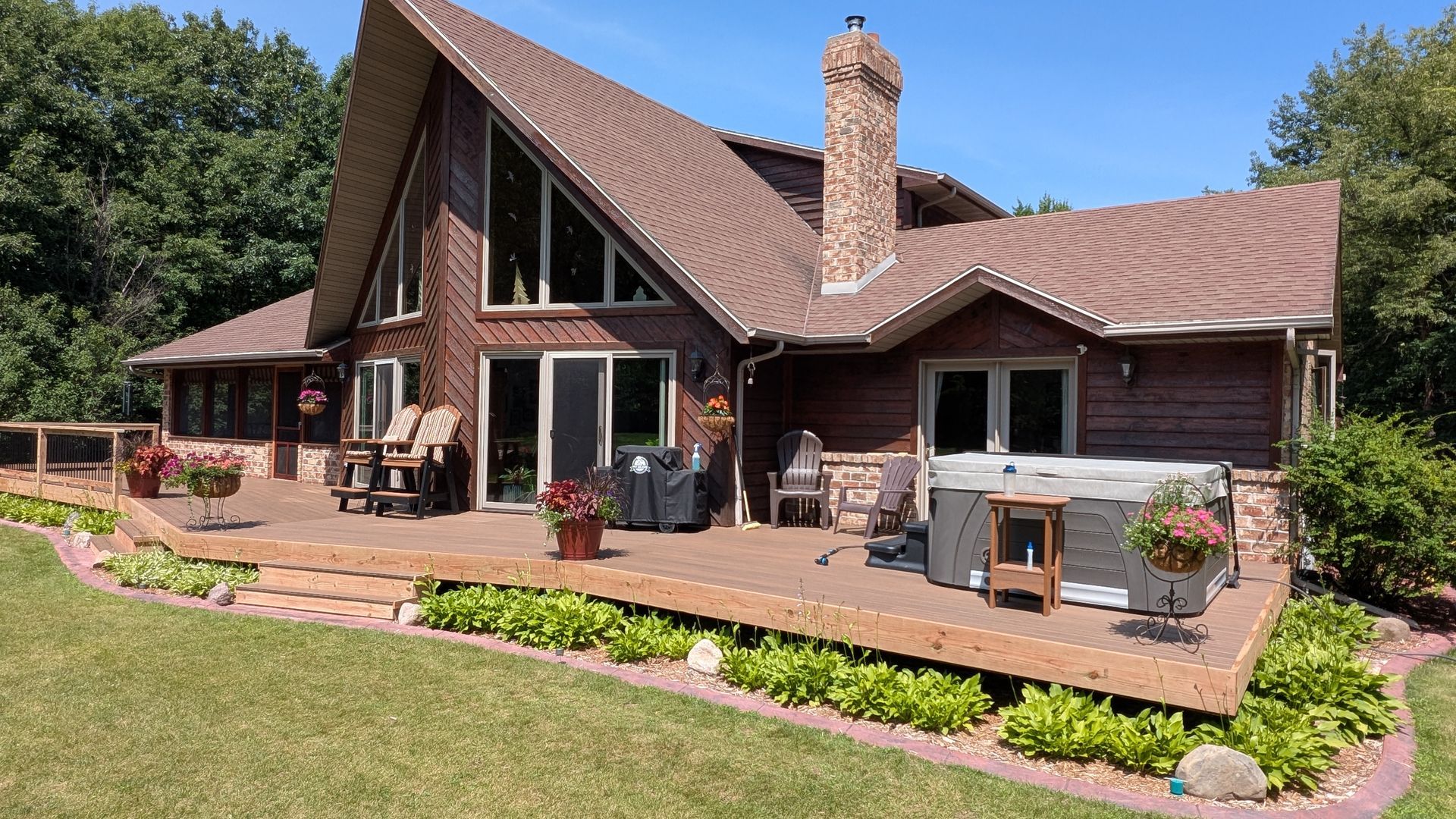 Rustic house with steep roof, brick chimney, wood deck, and flower beds in a sunny garden