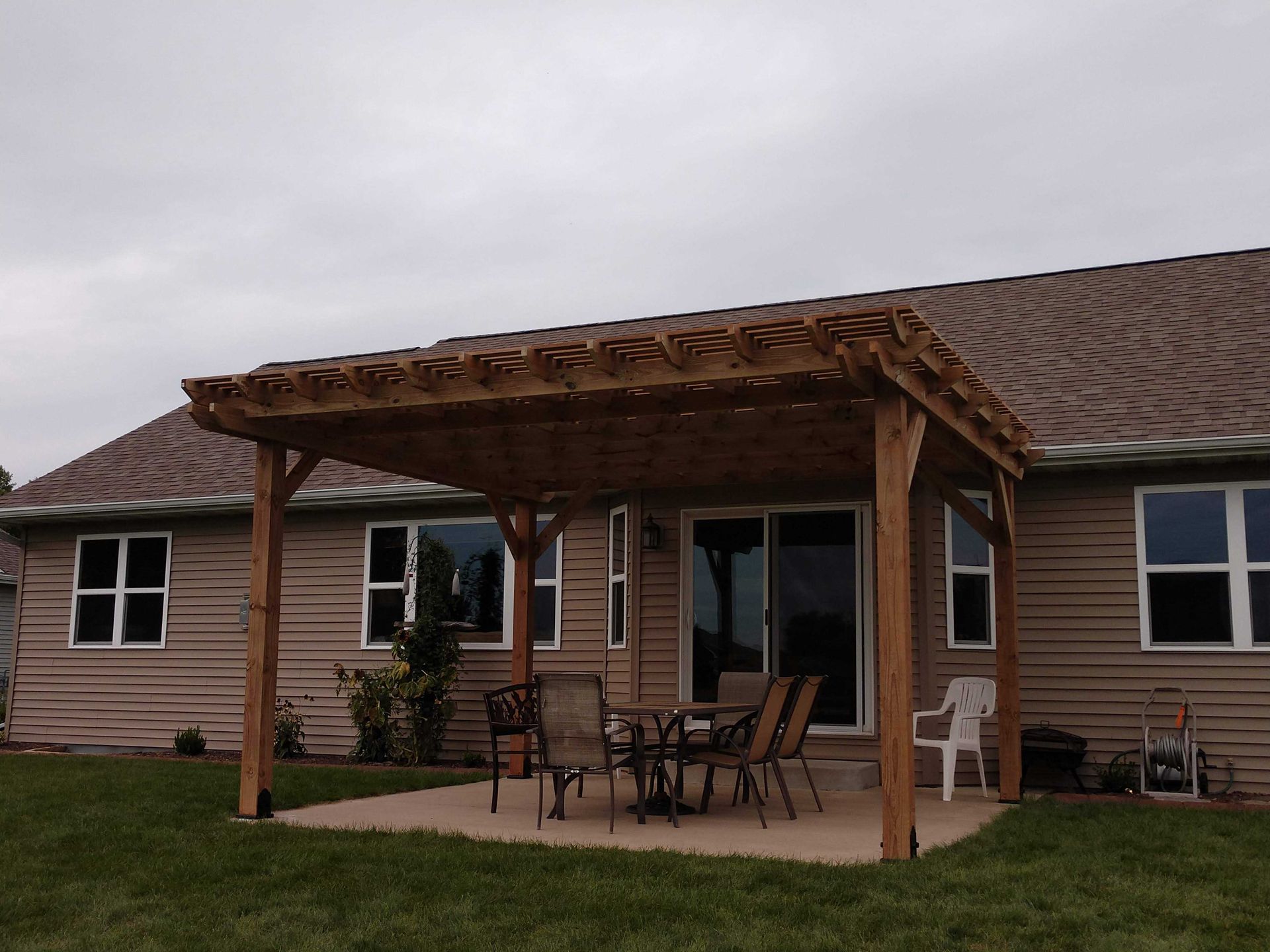 Wooden pergola over a patio with outdoor furniture, adjacent to a house with windows. Cloudy day.