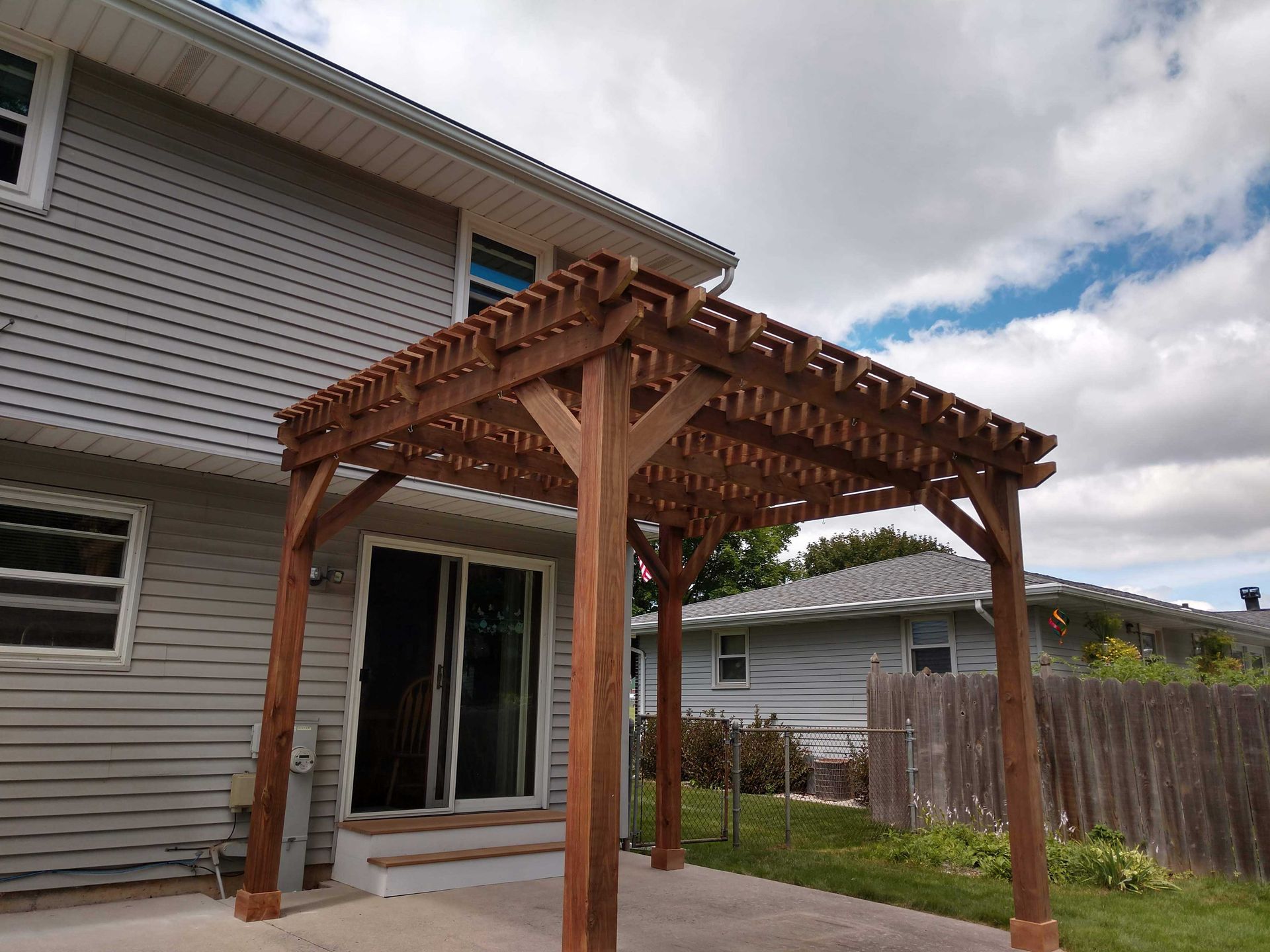 Brown wooden pergola attached to a gray house, over a patio. Cloudy sky in the background.