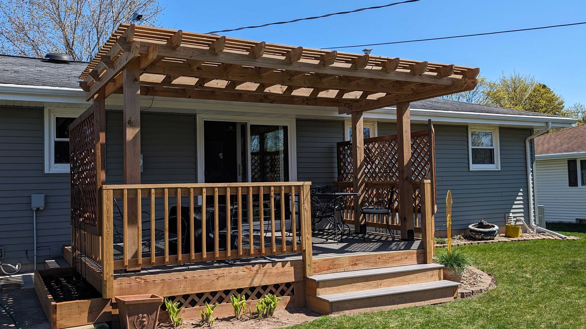 Wooden deck with pergola attached to a gray house, with seating and decorative lattice panels.