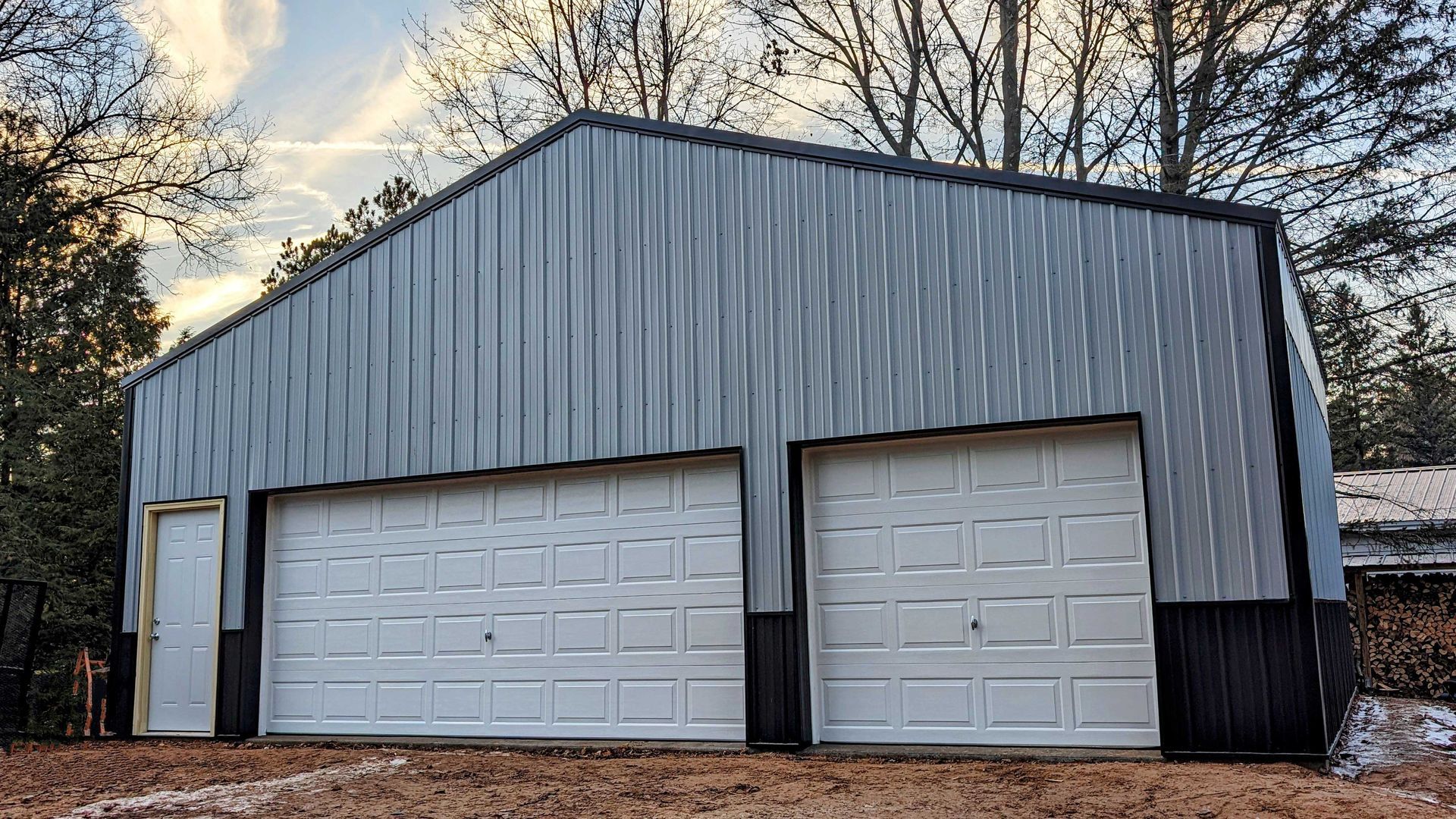 Two-bay metal garage with white doors, gray siding, and a black trim. Set in a wooded area.