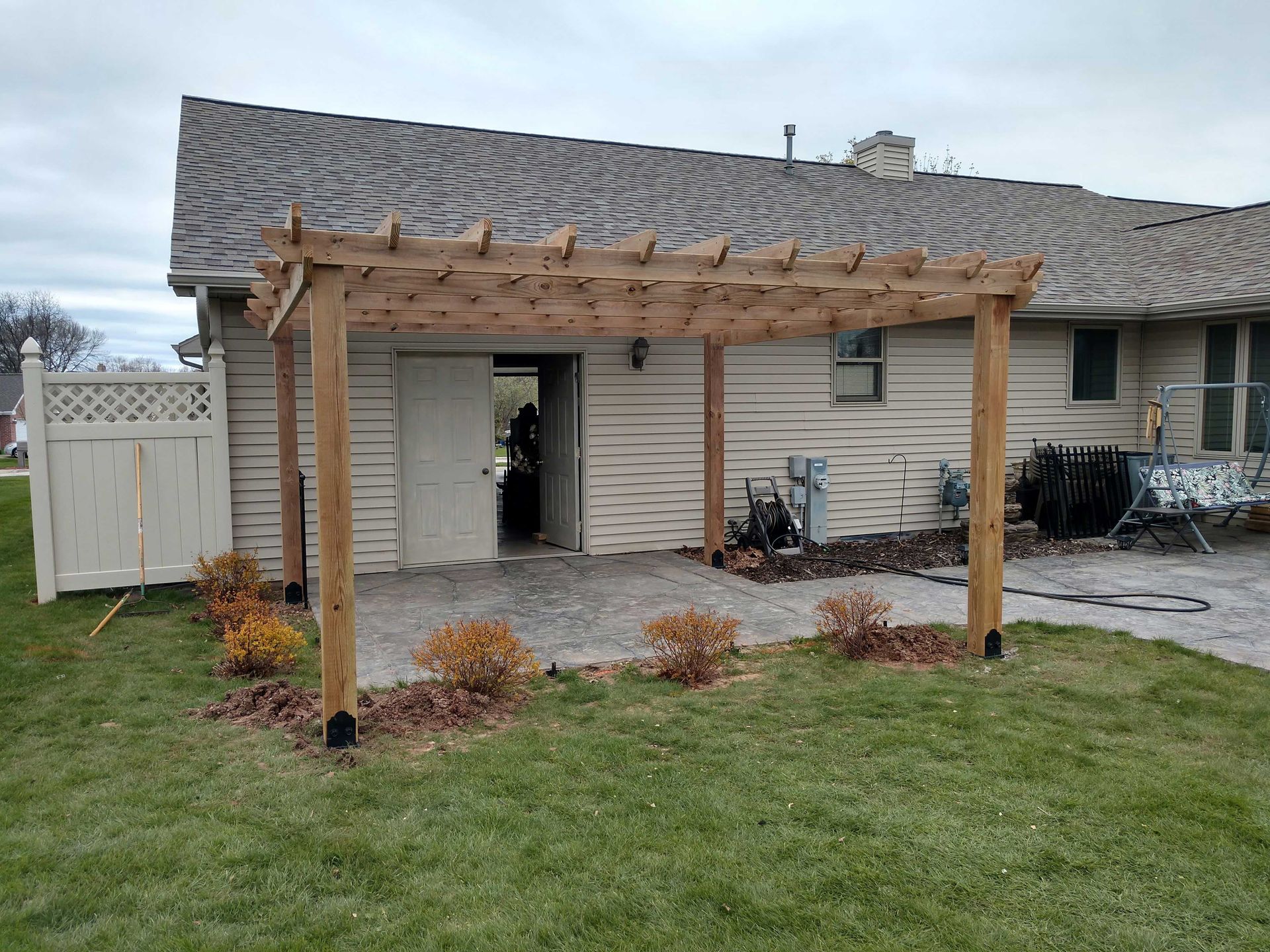 Wooden pergola over a concrete patio outside a house. Green grass and small bushes surround it.