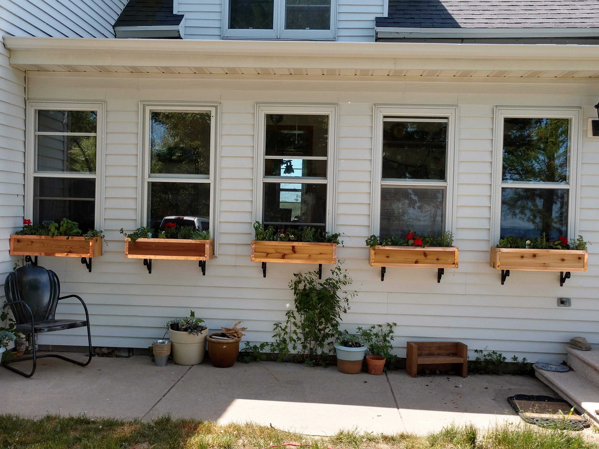 White house with five windows, each with a wooden window box and plants. A chair and potted plants sit on the porch.