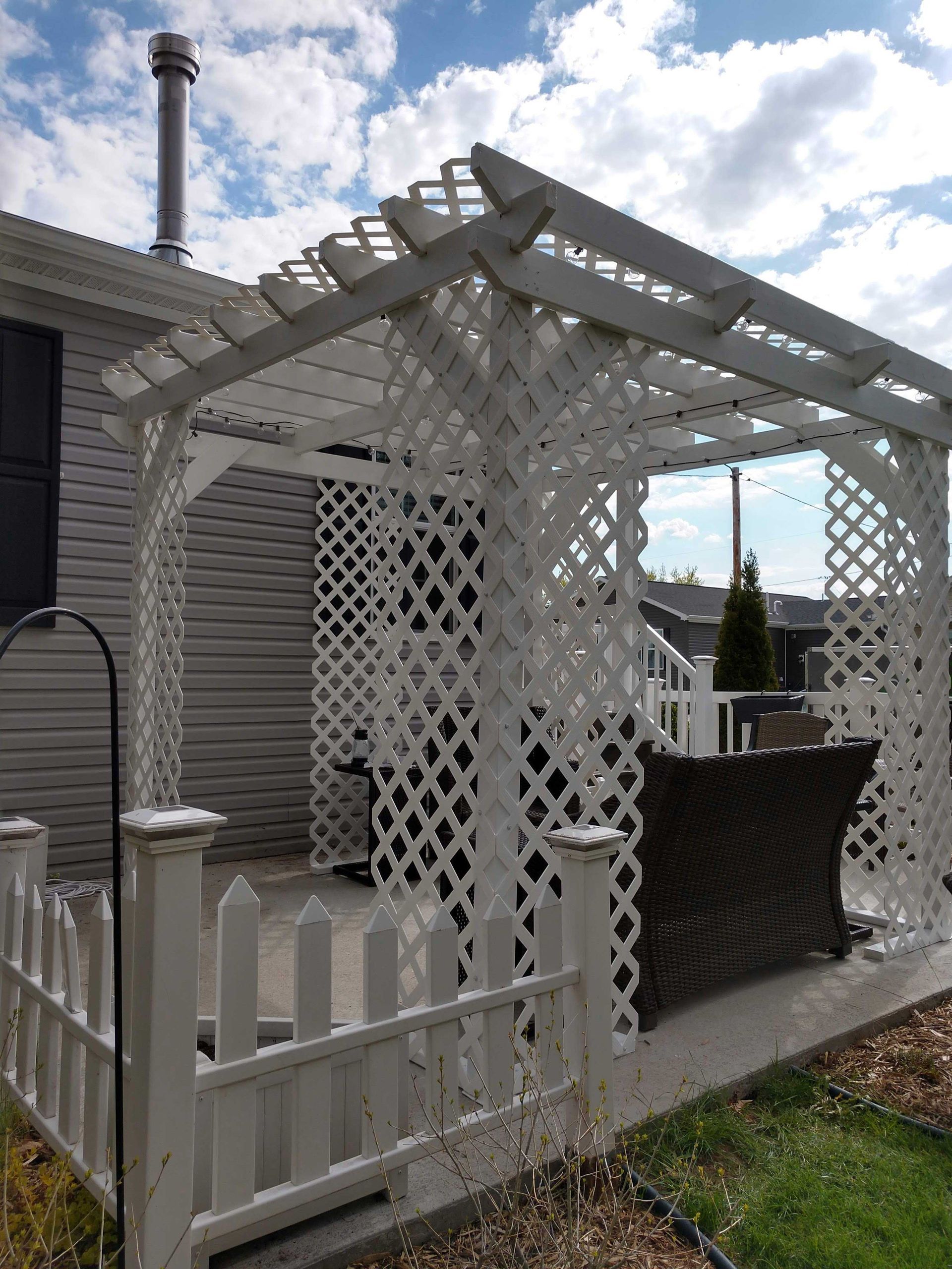 White lattice pergola with a matching fence, next to a house under a blue sky.