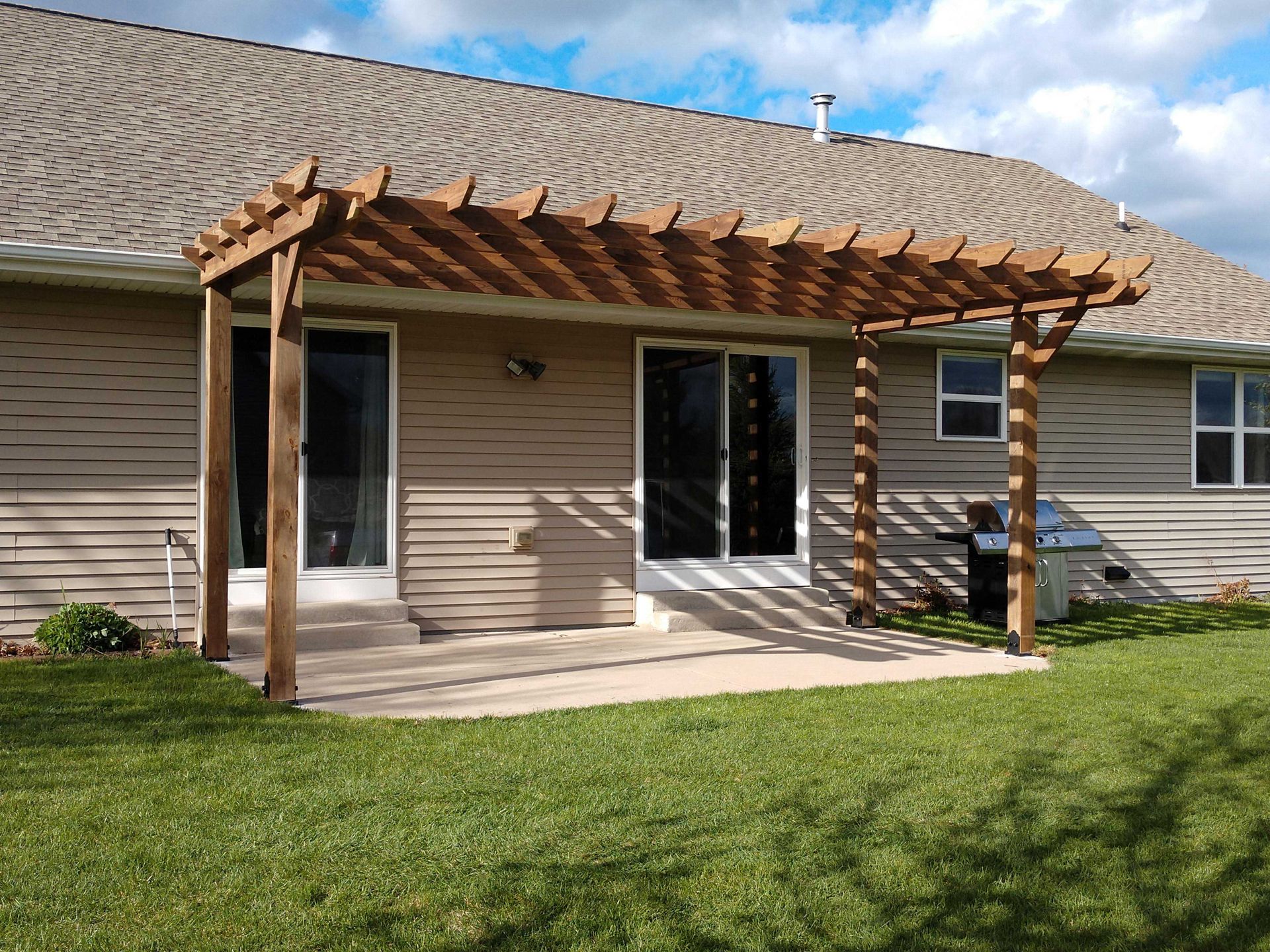 Wooden pergola over a concrete patio outside a house with sliding glass doors; green lawn.