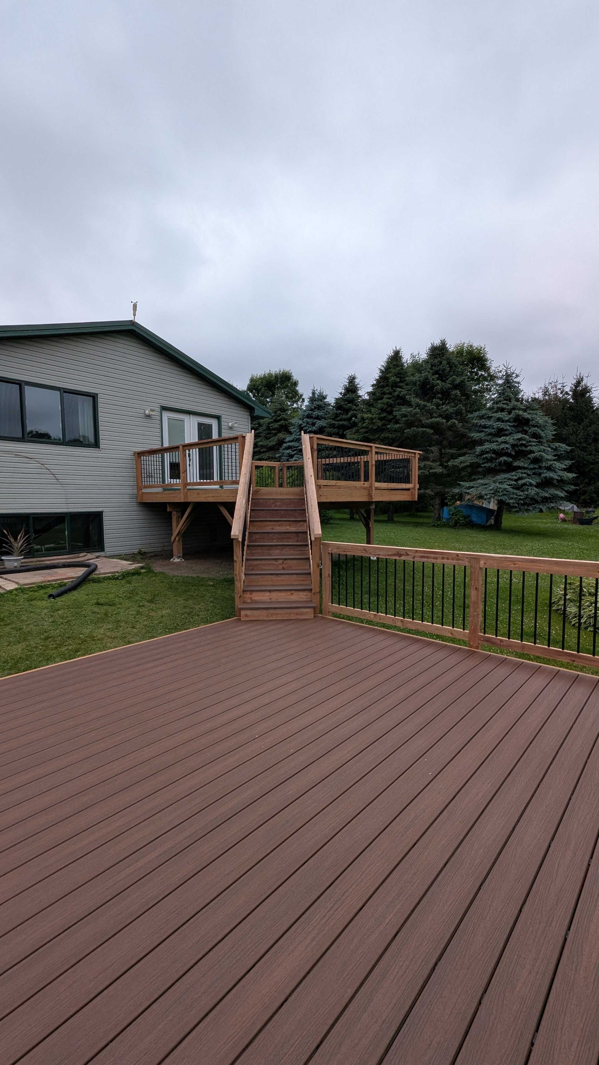 A wooden deck with stairs leads to a house with a door, surrounded by green grass and trees under a cloudy sky.