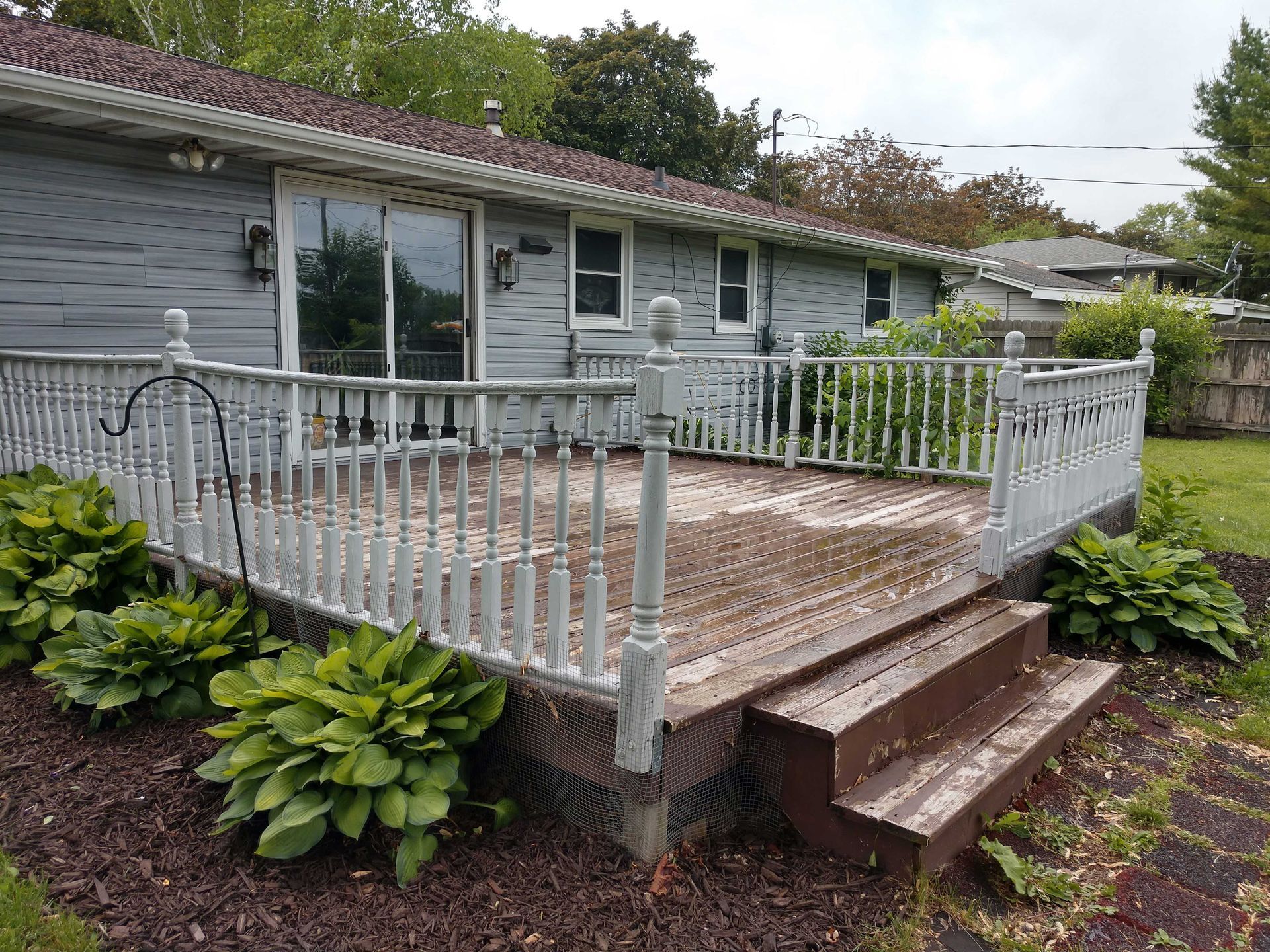 Grey house with a weathered wooden deck. Hosta plants surround the deck. Brown mulch is visible.
