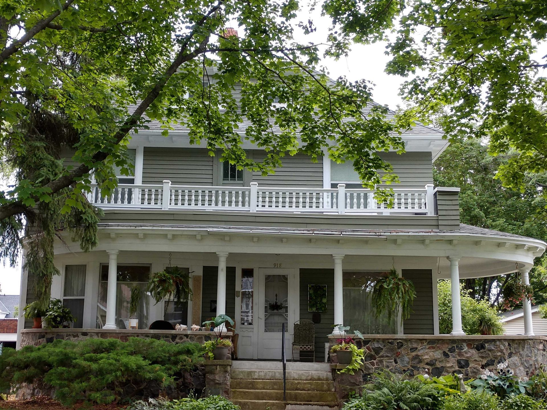 Two-story house with a wraparound porch and second-floor balcony, surrounded by trees.