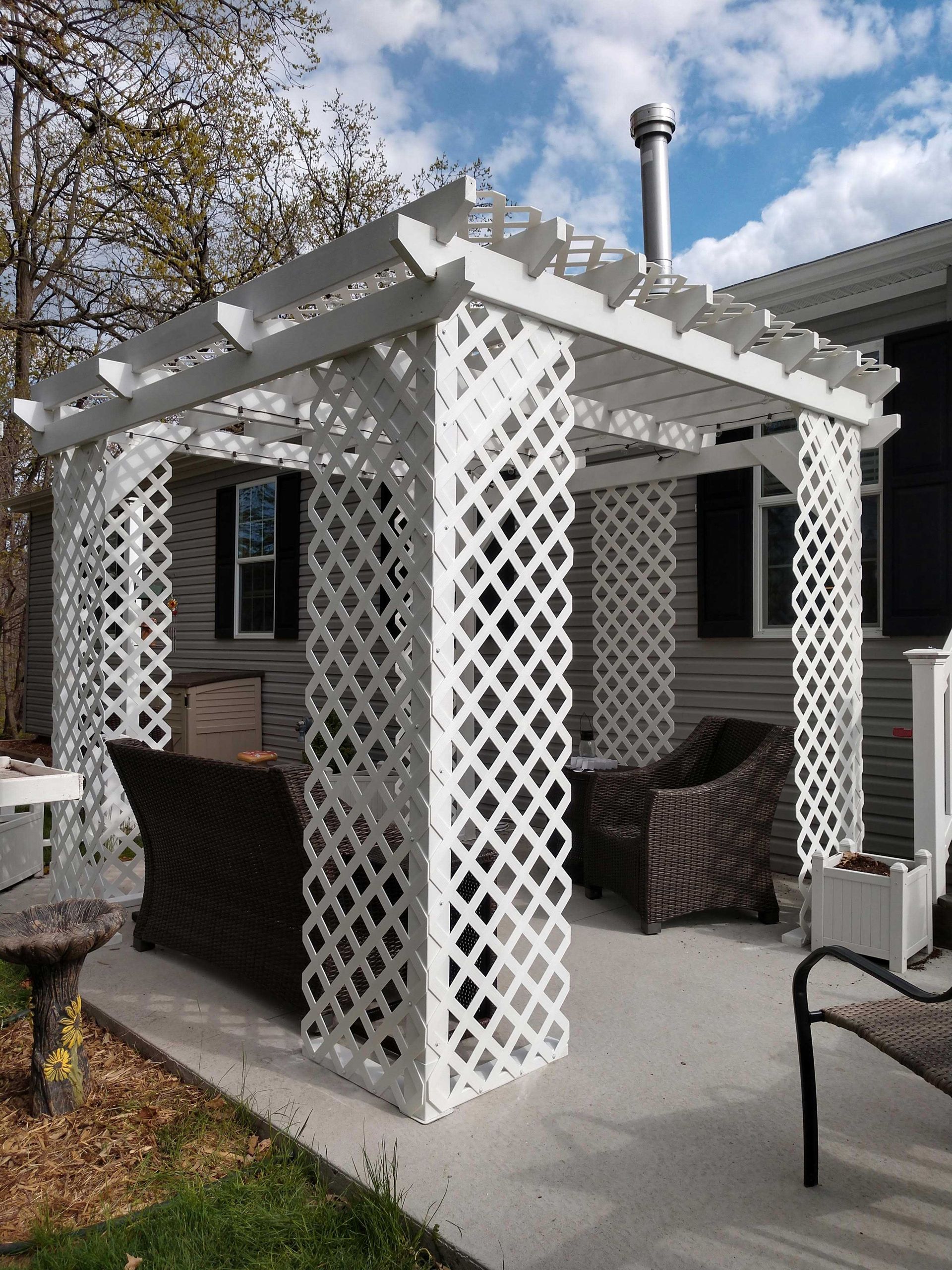 White lattice pergola with seating area on a concrete patio next to a building.