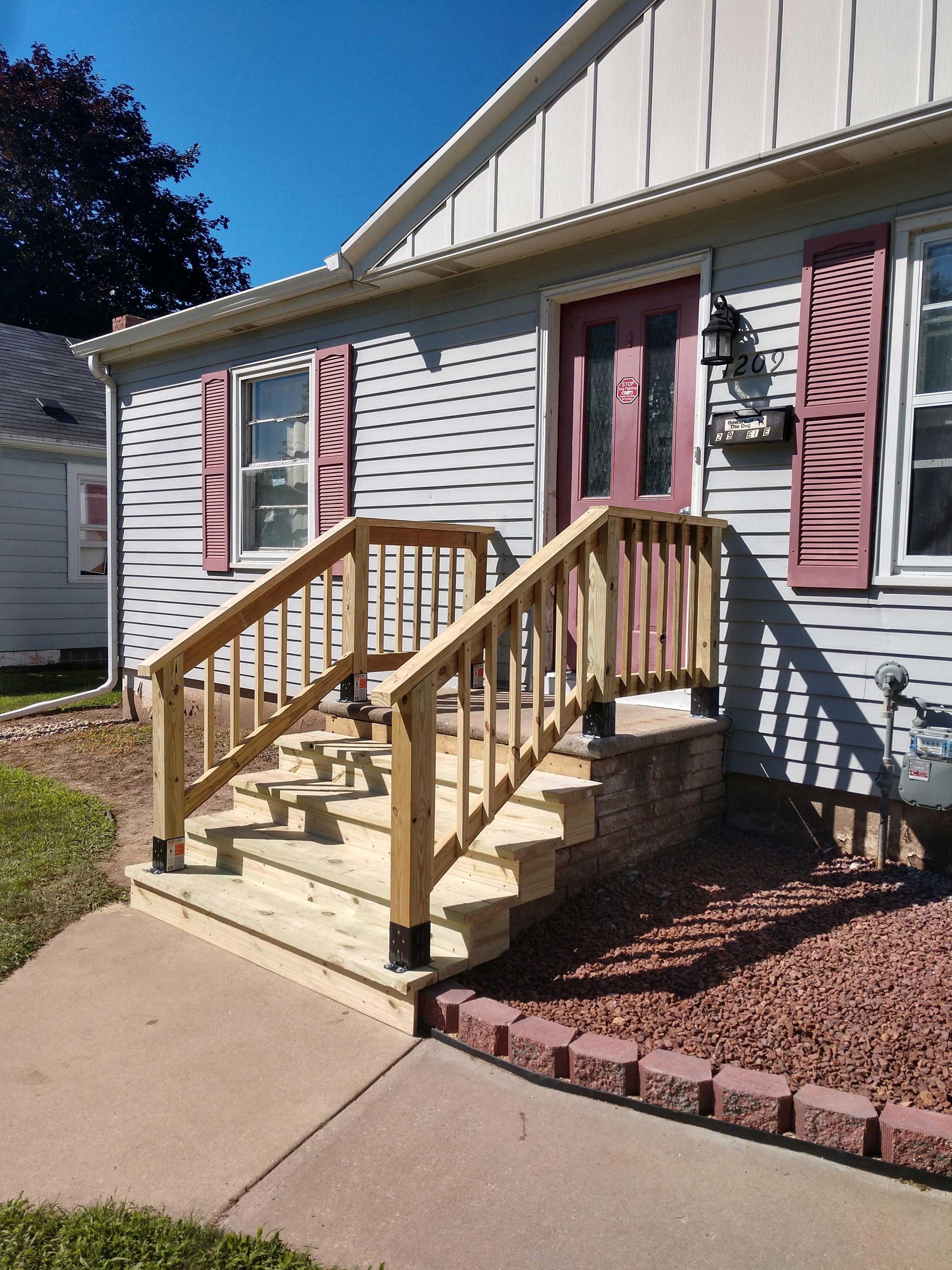 A wooden staircase leads to a light blue house with a red door. Red shutters flank windows.