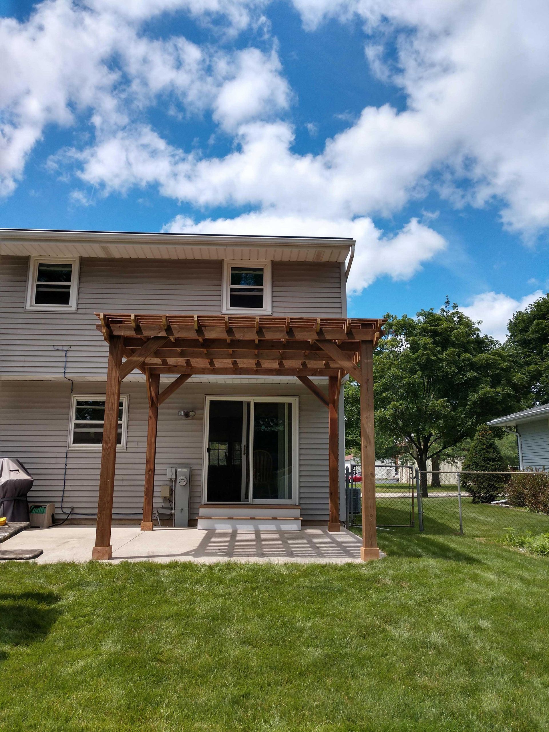 Wooden pergola attached to a two-story house with a sliding glass door. Overcast sky, green grass.