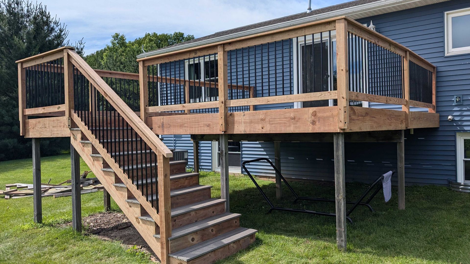 Wooden deck with stairs attached to a blue house, set on green grass. Black railings present.