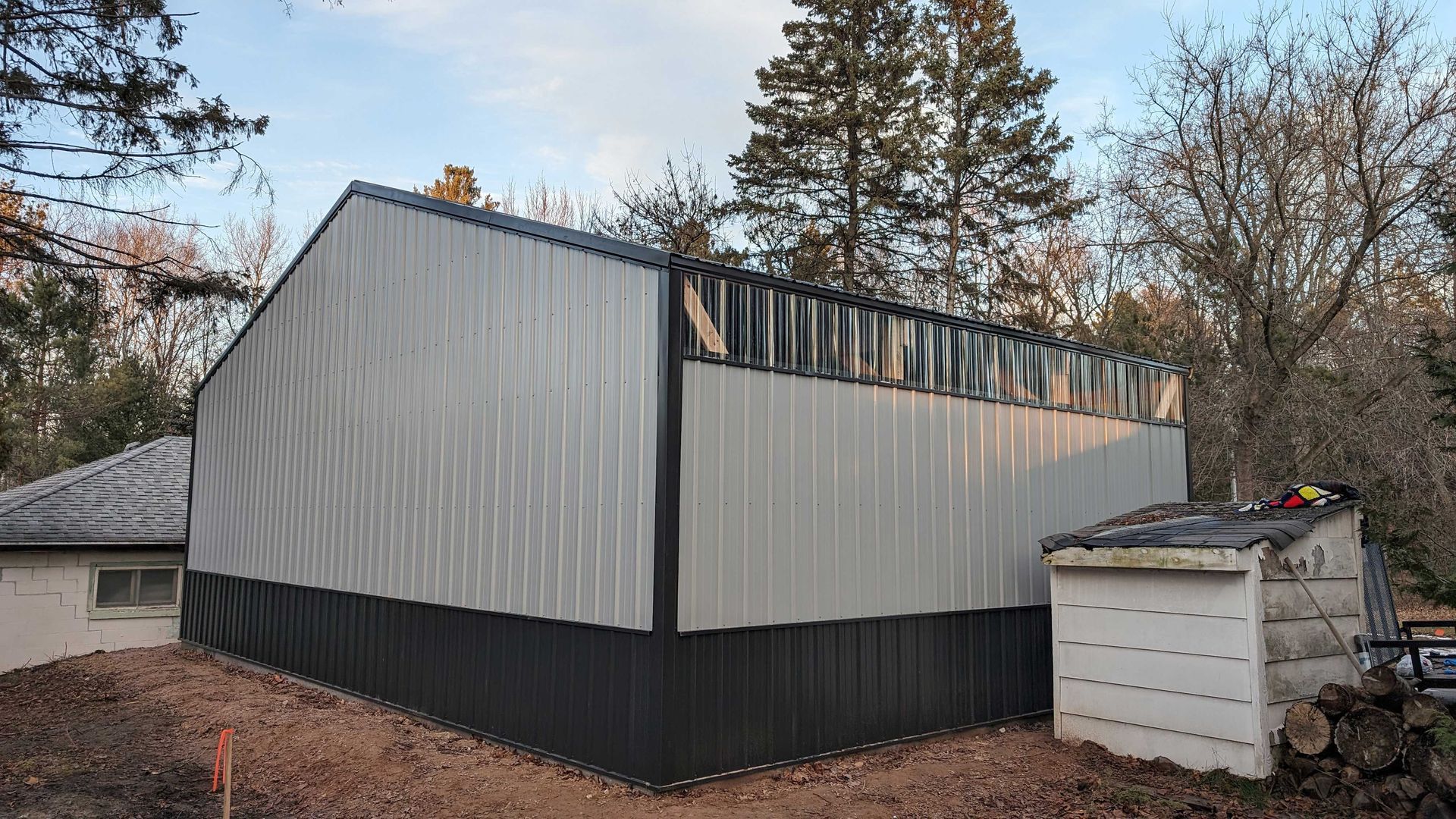 Metal-sided building under construction, grey and black panels, with a smaller building to the right. Bare trees in background.