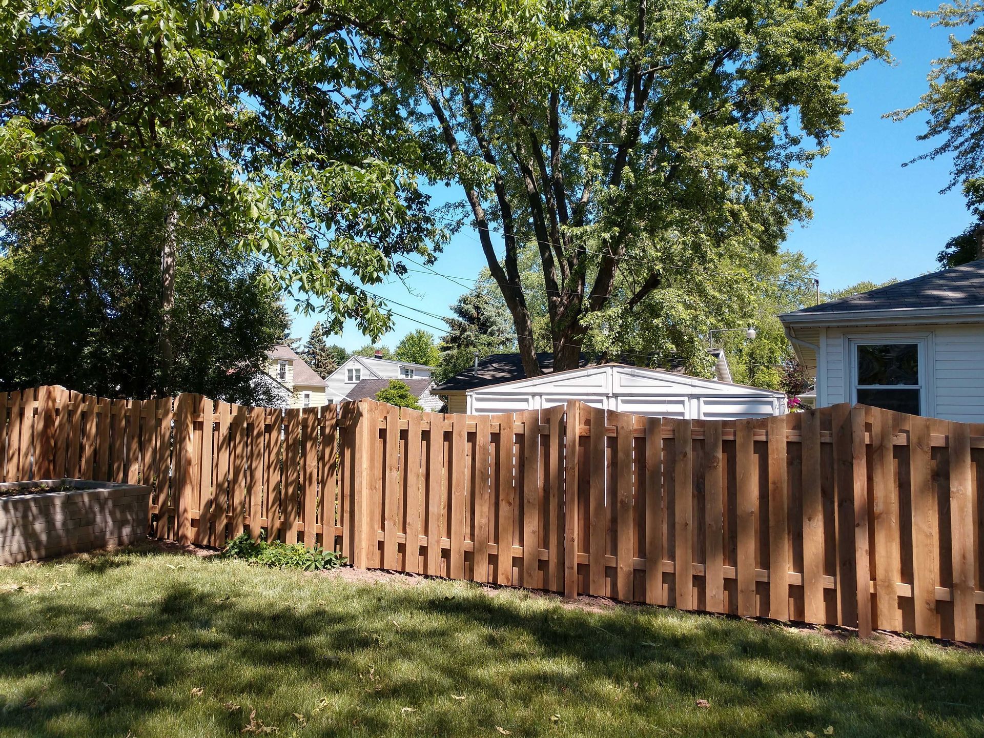 Brown wooden fence in a grassy backyard, trees and houses in the background under a blue sky.