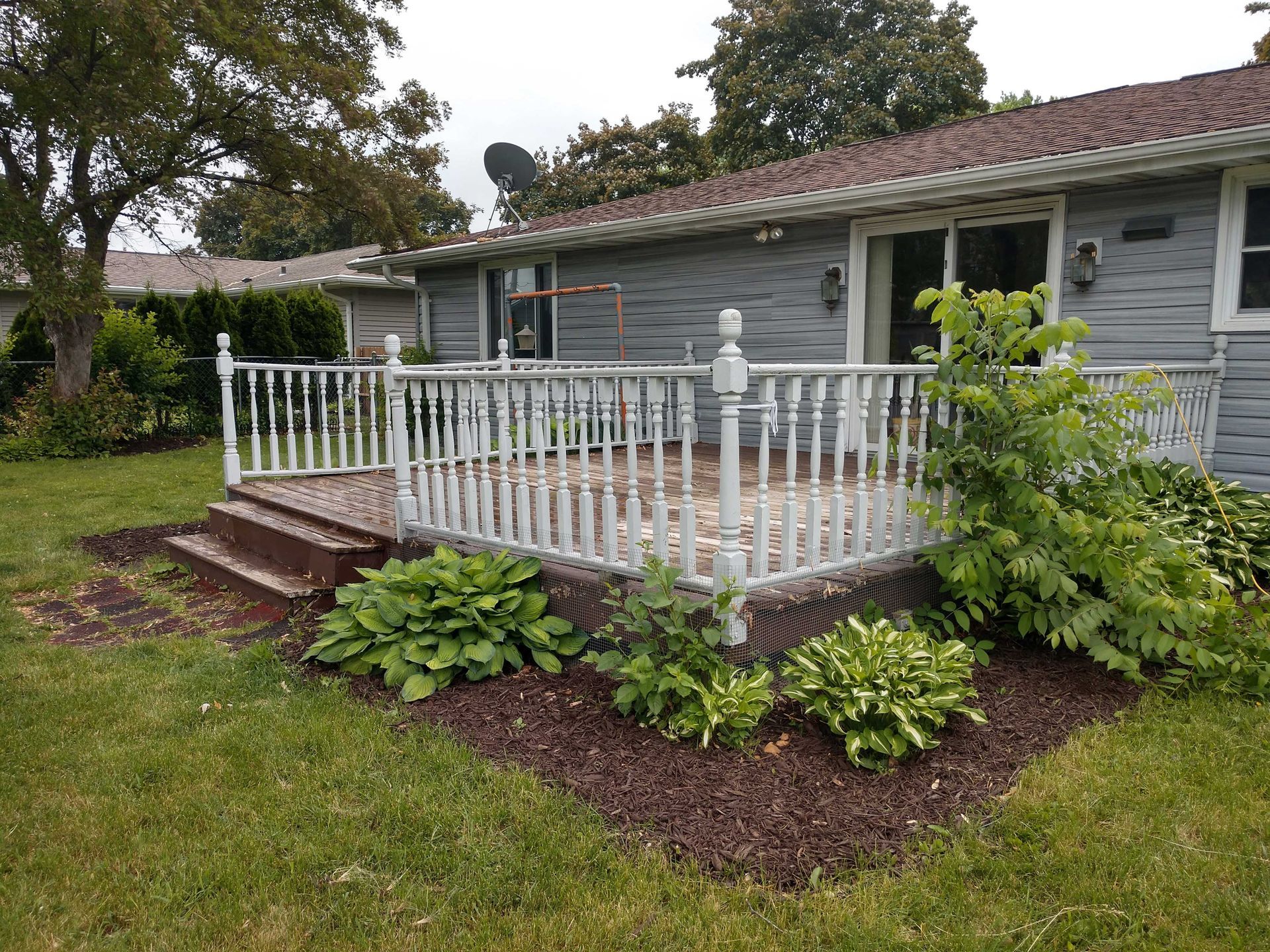 Deck with white railing and steps, against a gray house, surrounded by mulch and greenery.