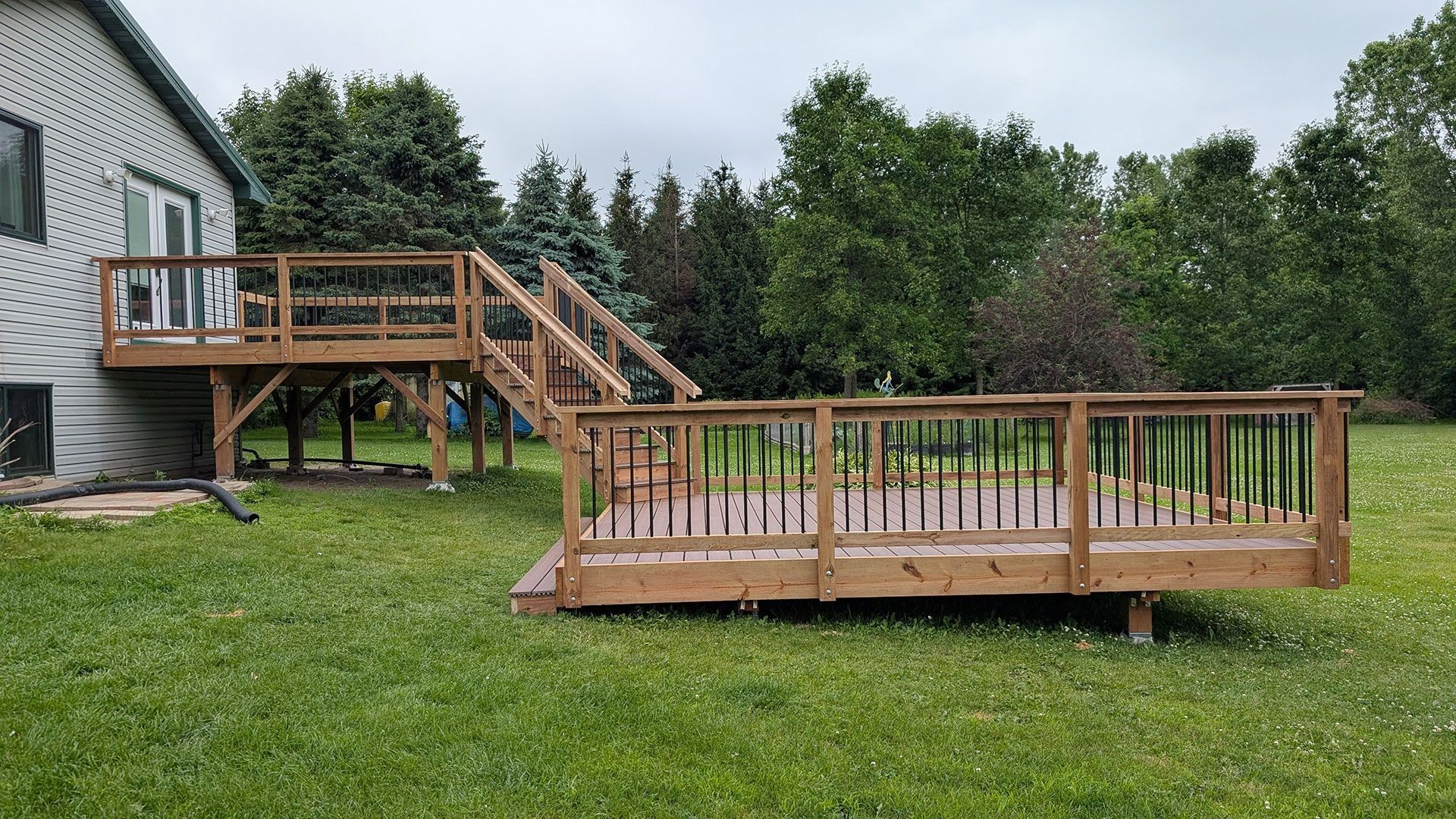 A two-level wooden deck attached to the back of a house, set in a grassy backyard surrounded by mature trees.