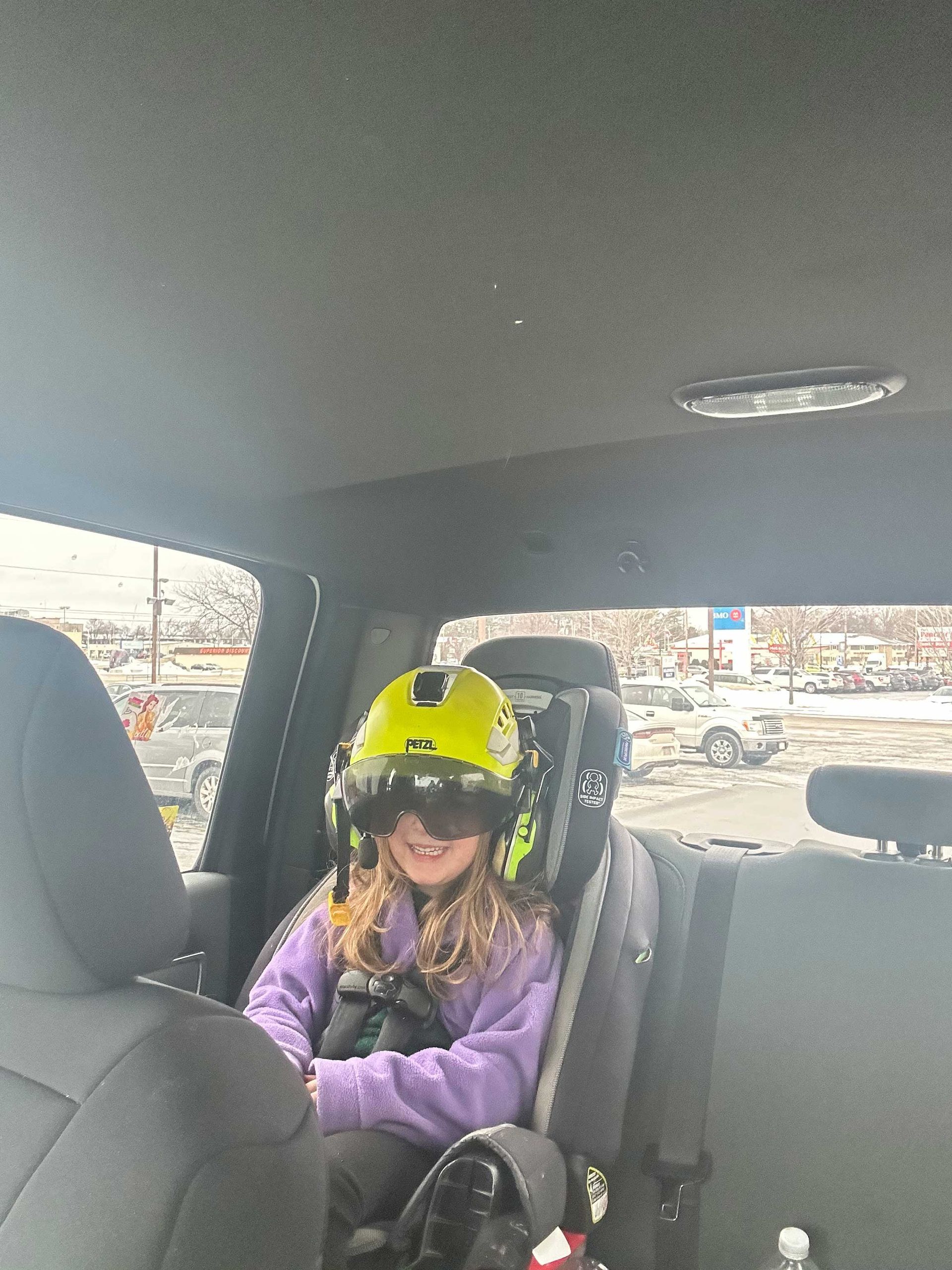 Girl in back seat wearing a yellow helmet and goggles; sitting in a truck with a backpack next to her.