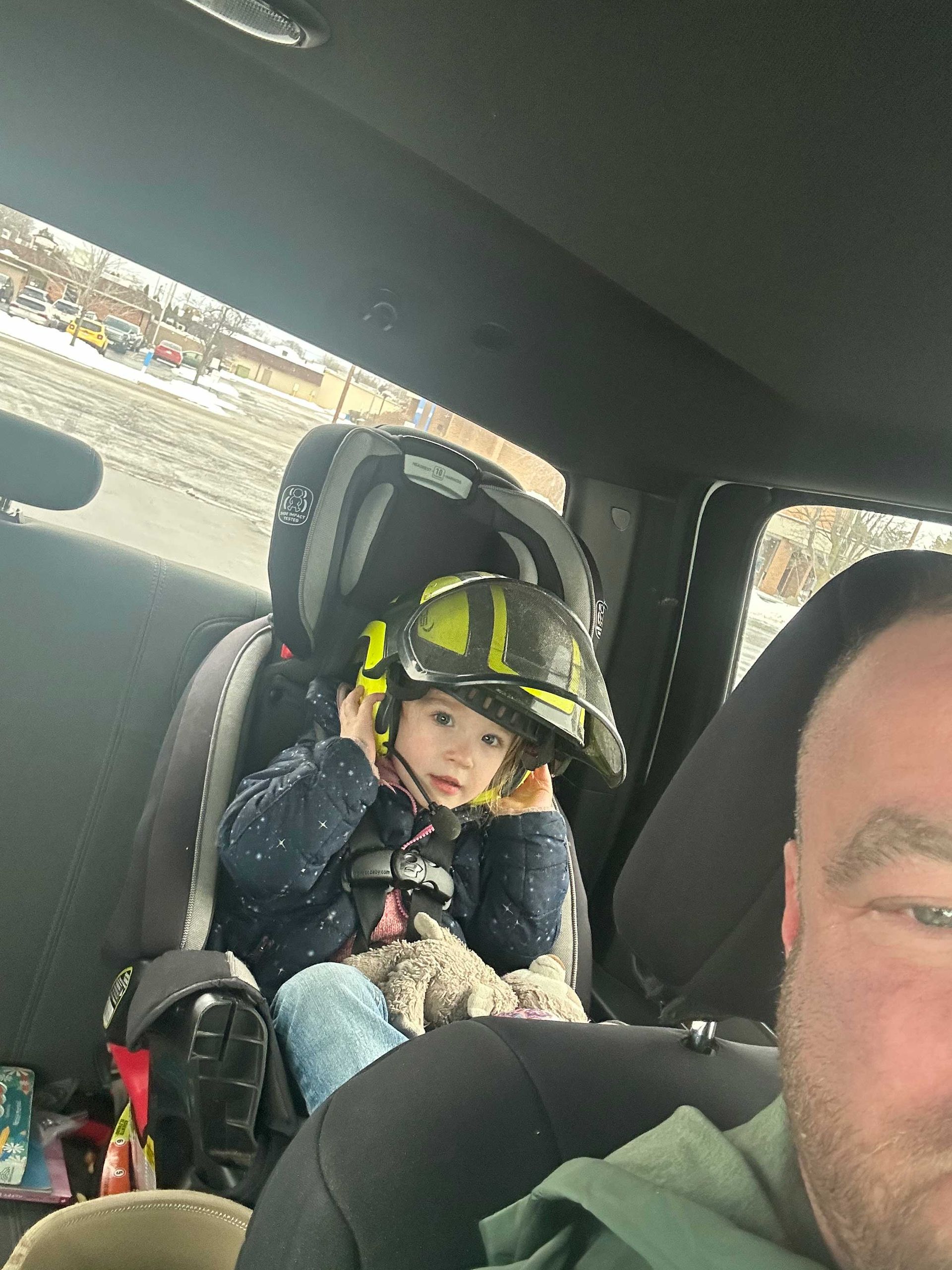 Child in car seat wearing a firefighter helmet, next to an adult.