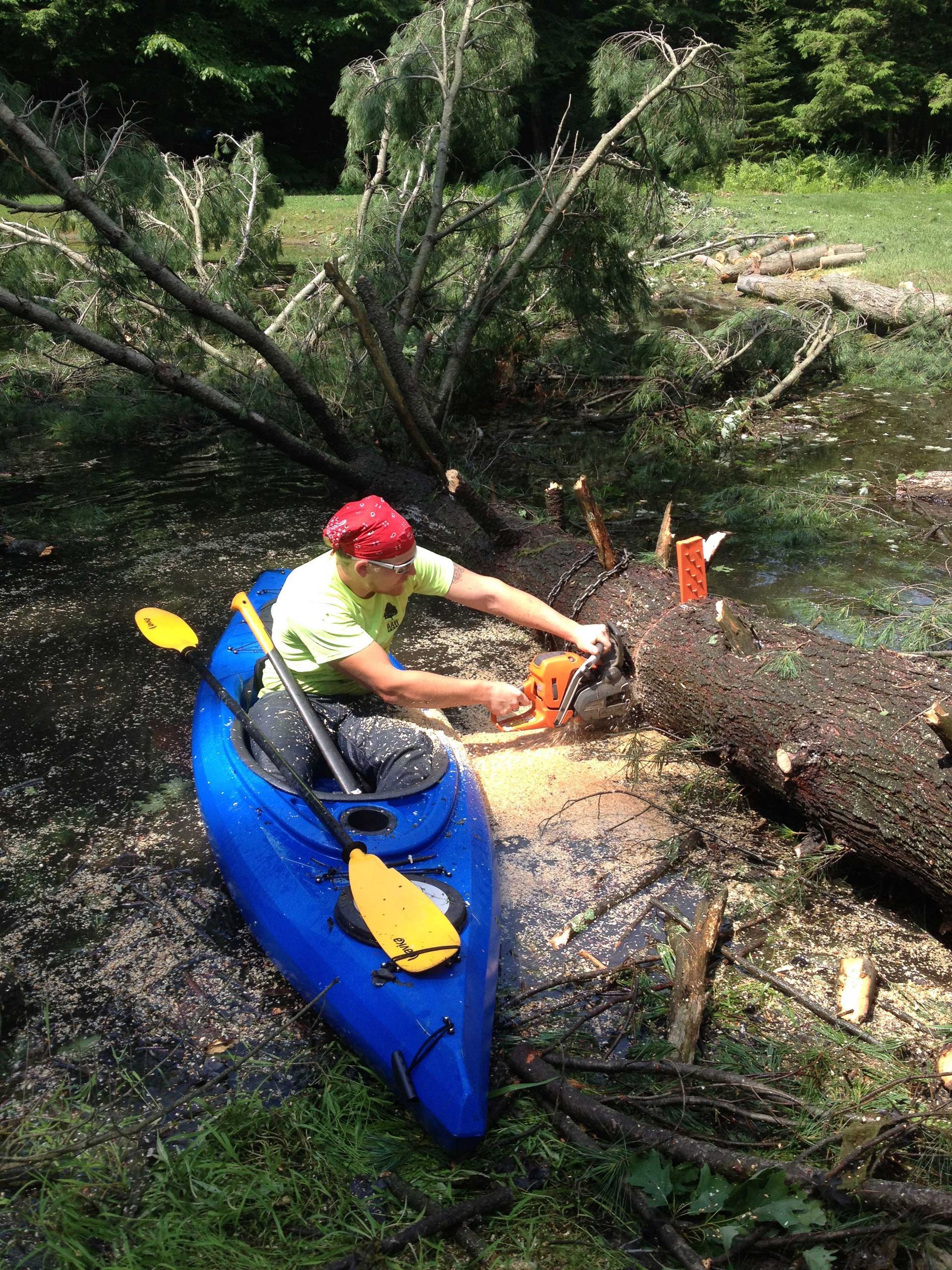 Man in blue kayak uses chainsaw on fallen tree in water.