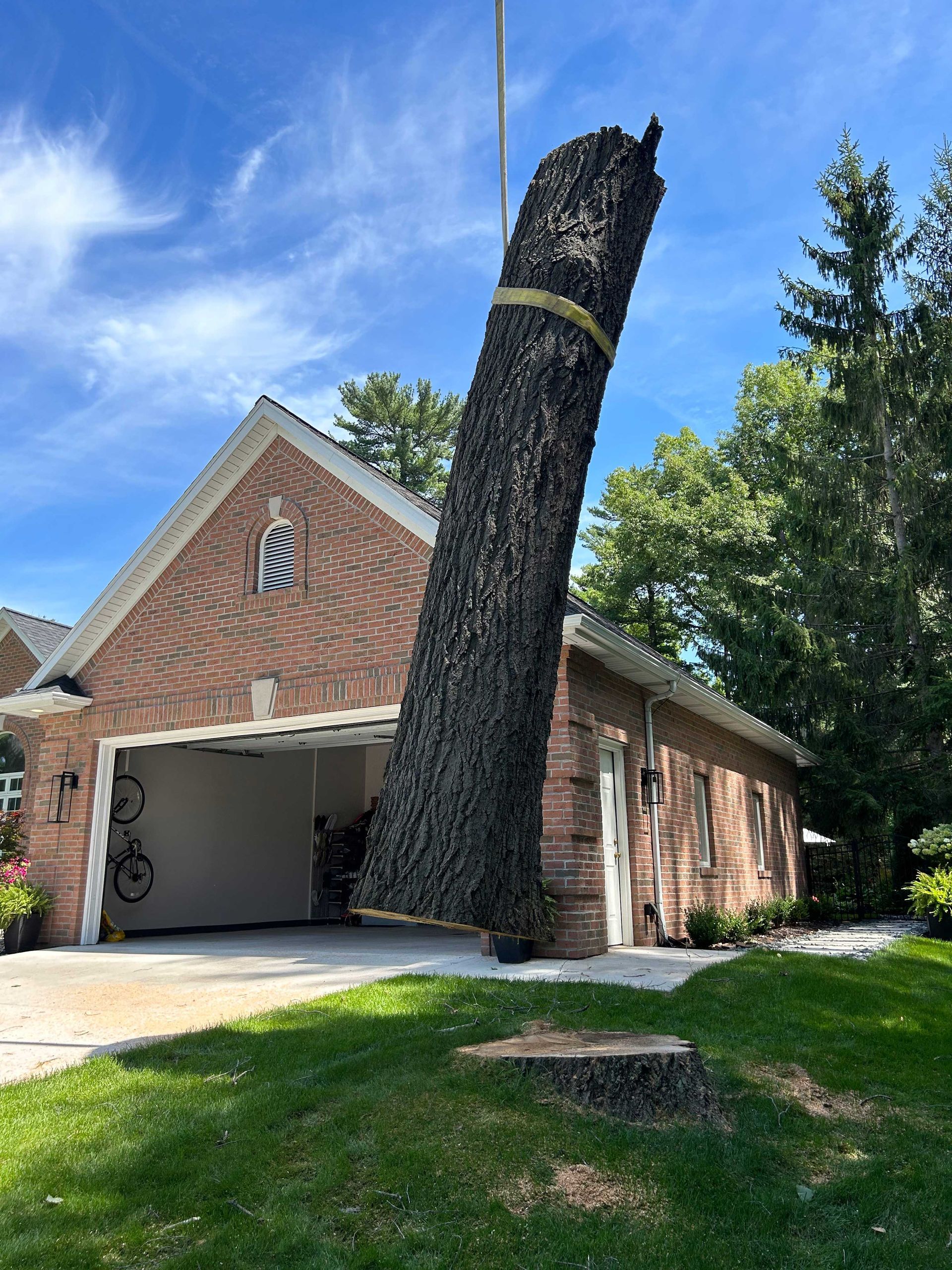 A long tree trunk is lifted by a crane near a brick house with an open garage on a sunny day.