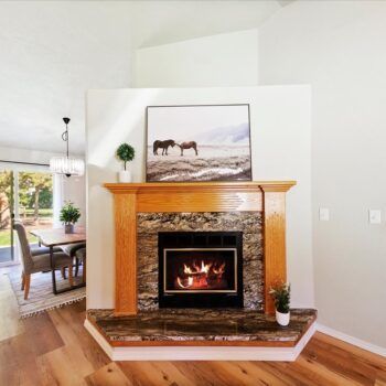 A corner fireplace with a dark marble surround, light oak wood mantel, and framed horse print, next to a dining area.