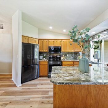 A kitchen with honey-oak cabinets, dark granite countertops, a black refrigerator, and light wood-plank floors.