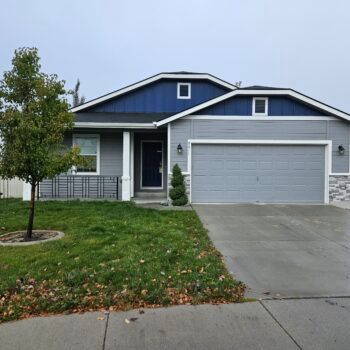 A one-story suburban home with blue and grey siding, a two-car garage, stone accents, and a young tree in the front lawn.