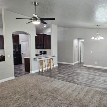 A view of an open-concept living room with gray carpet and wood-look floors, a kitchen counter with stools, and cabinets.
