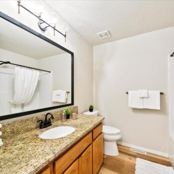 A bathroom with a granite-topped vanity, a large framed mirror, a toilet, and white towels on a wall rack.